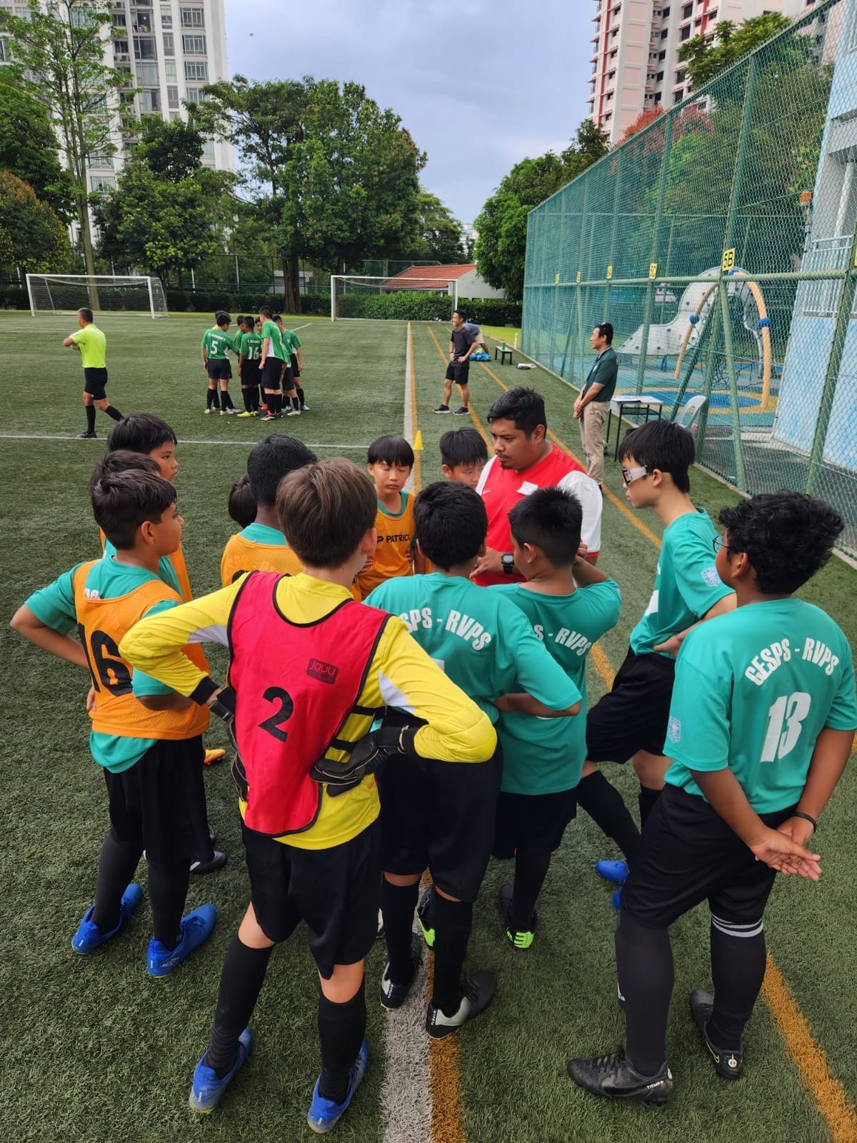 Young soccer players in teal GESPS-RVPS jerseys huddle with a coach on a green field during a match.
