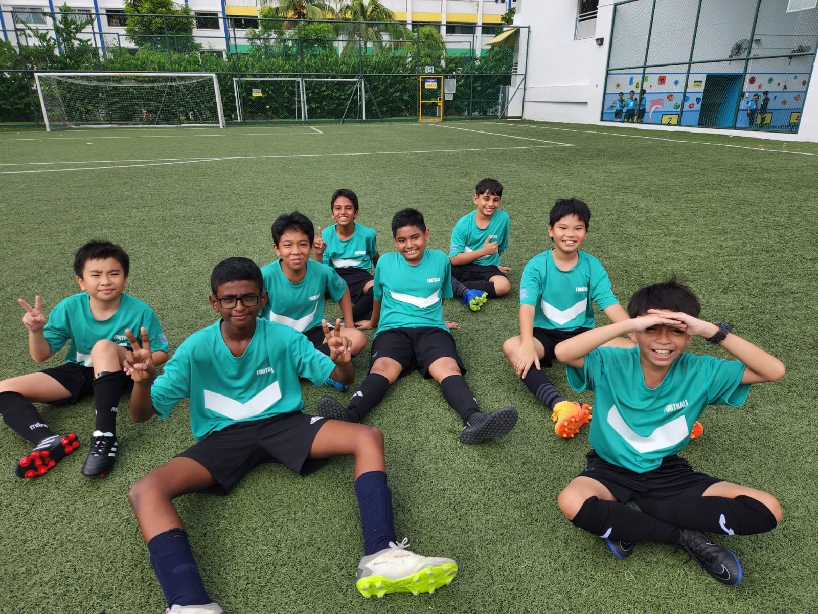 Eight boys in teal football jerseys and black shorts sit on a green artificial pitch, smiling and posing.