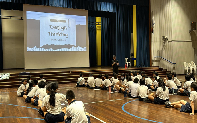 Students in uniform sit on a gym floor facing a presenter near a screen showing "Design Thinking Student Leadership."