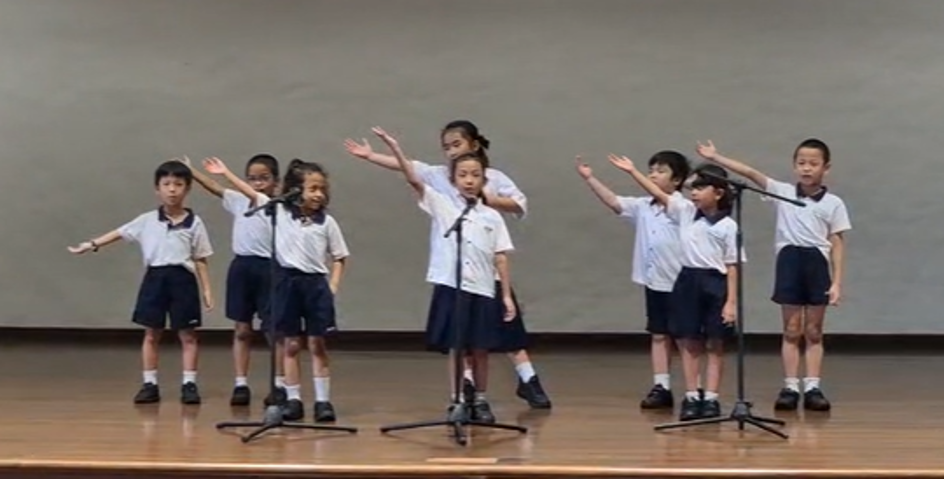 Eight young students in school uniforms perform on stage with microphones, arms outstretched in a choreographed routine.