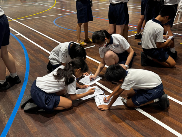 Students in white shirts and navy shorts kneel on a gym floor writing on papers together.