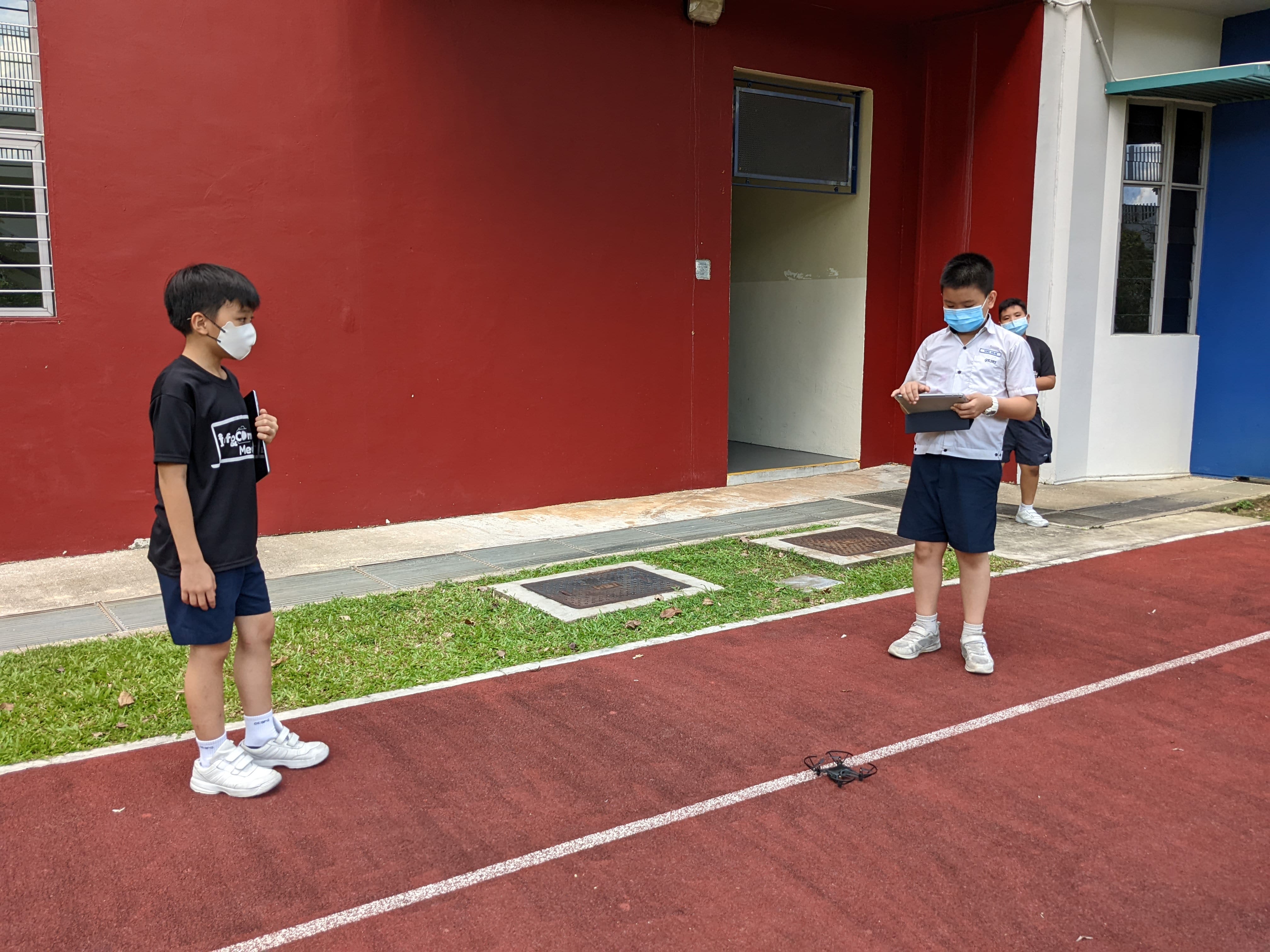 Two masked schoolboys stand on a red running track, one holding a tablet near a small drone on the ground.