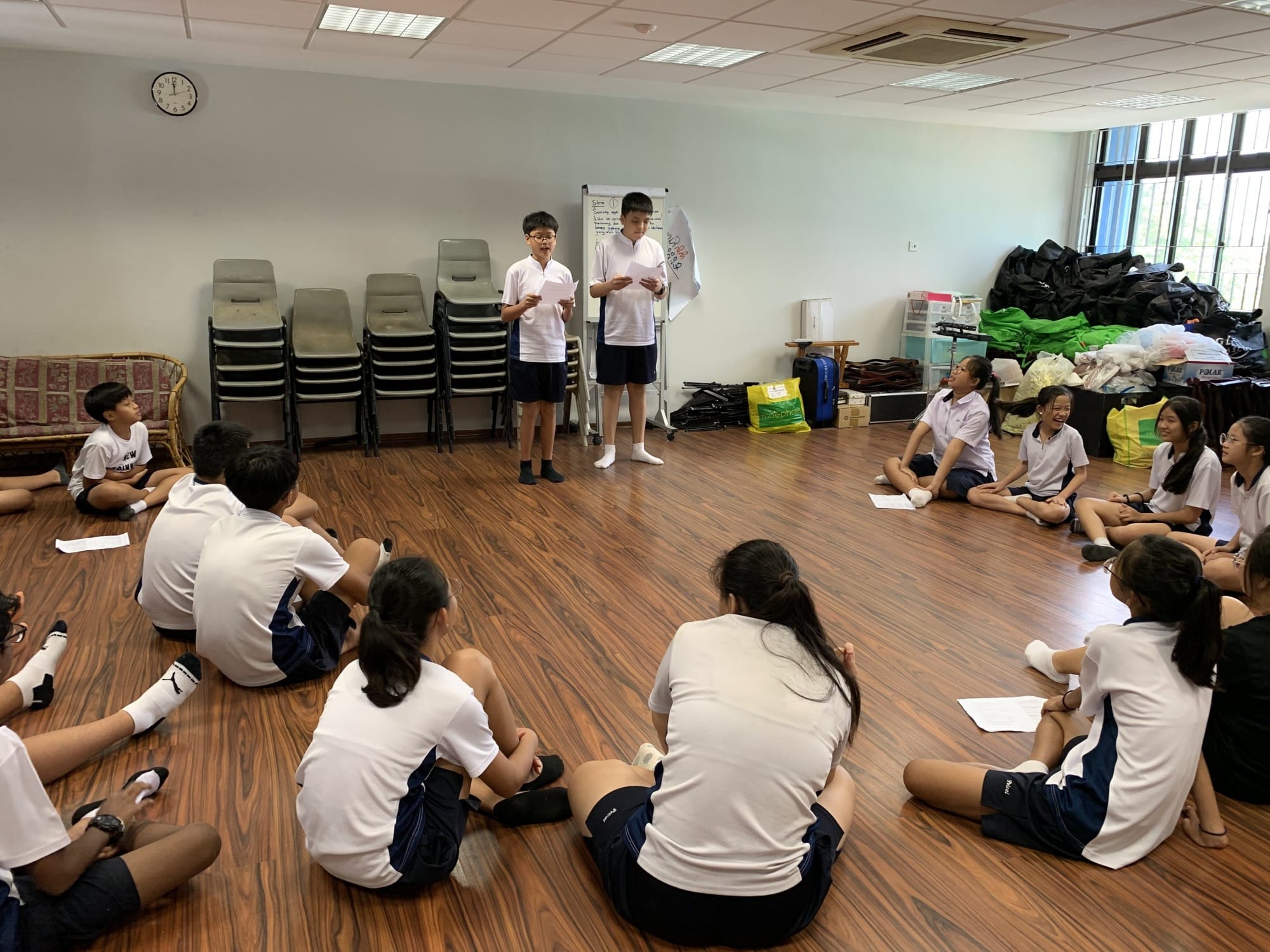 Two students in school uniforms stand reading papers while classmates sit in a circle on a wooden floor.