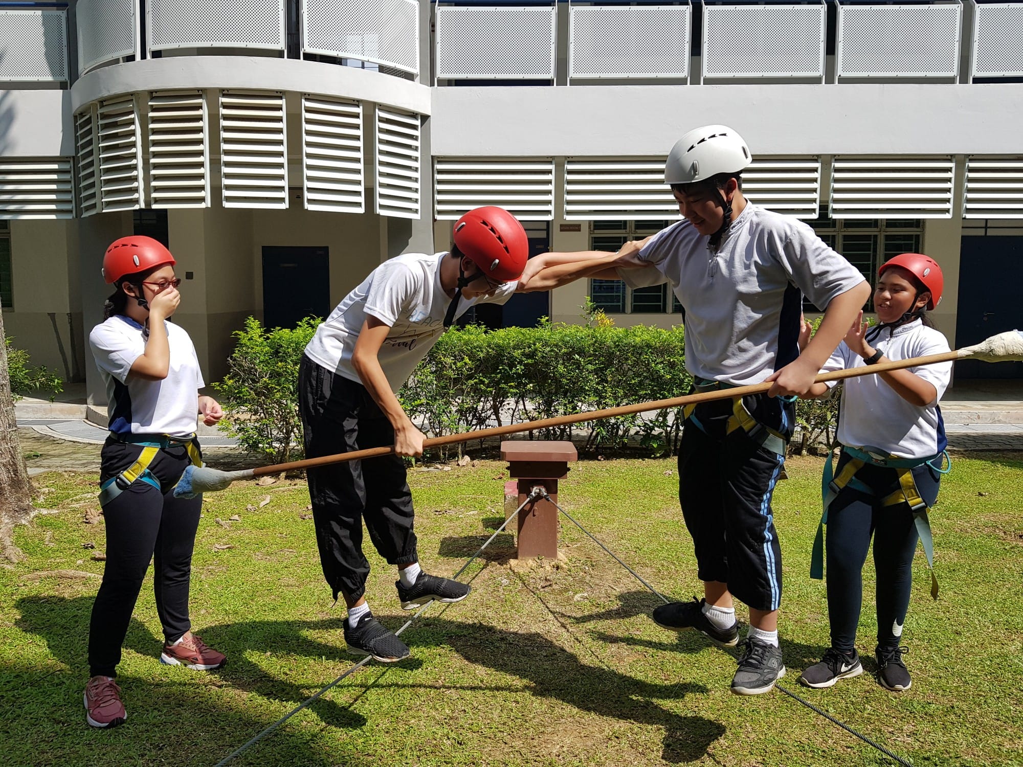 Four youths wearing helmets and harnesses balance on a rope course outdoors, holding wooden poles for support.