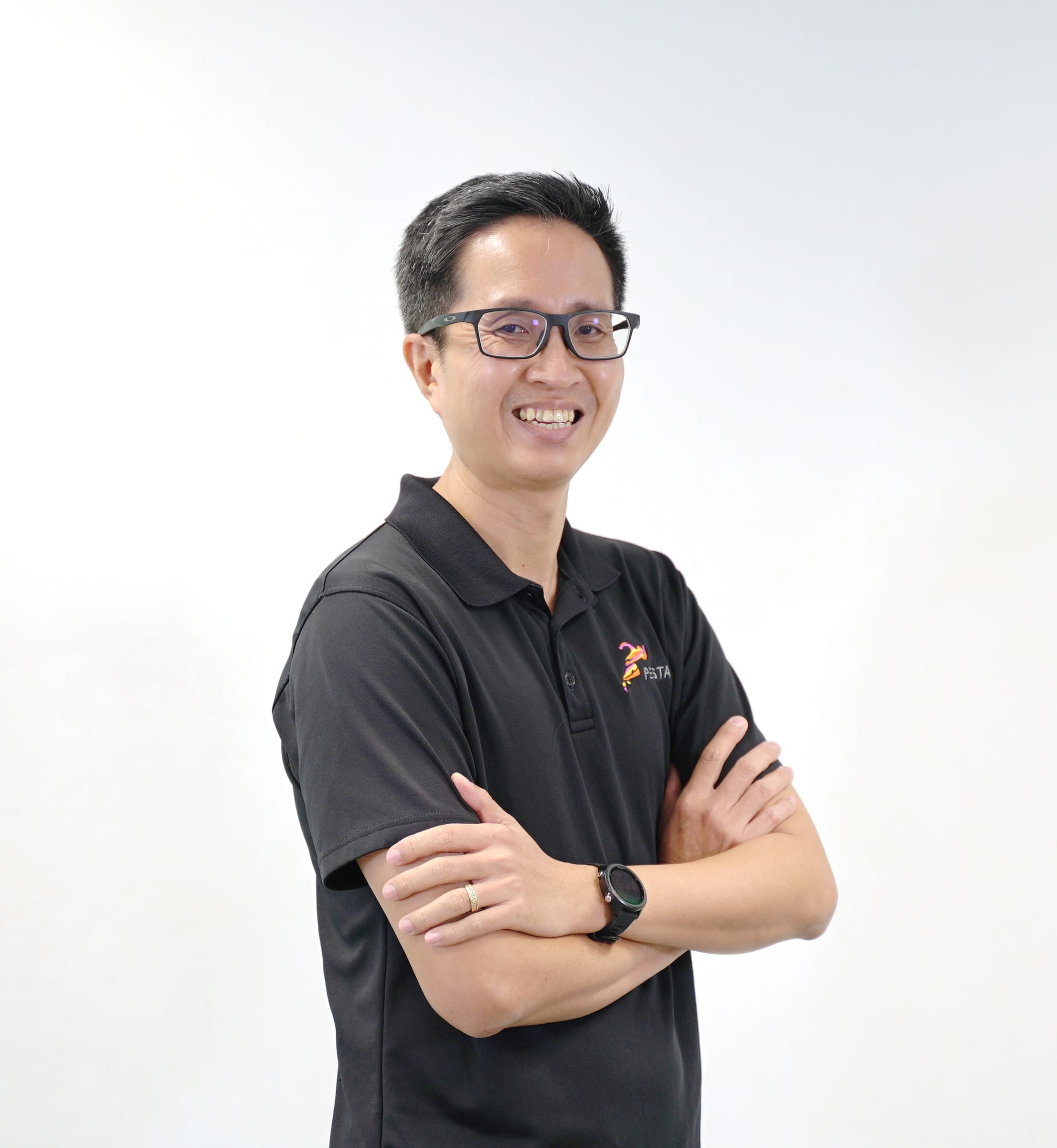 Smiling man with glasses and crossed arms wearing a black polo shirt with a logo, against a white background.