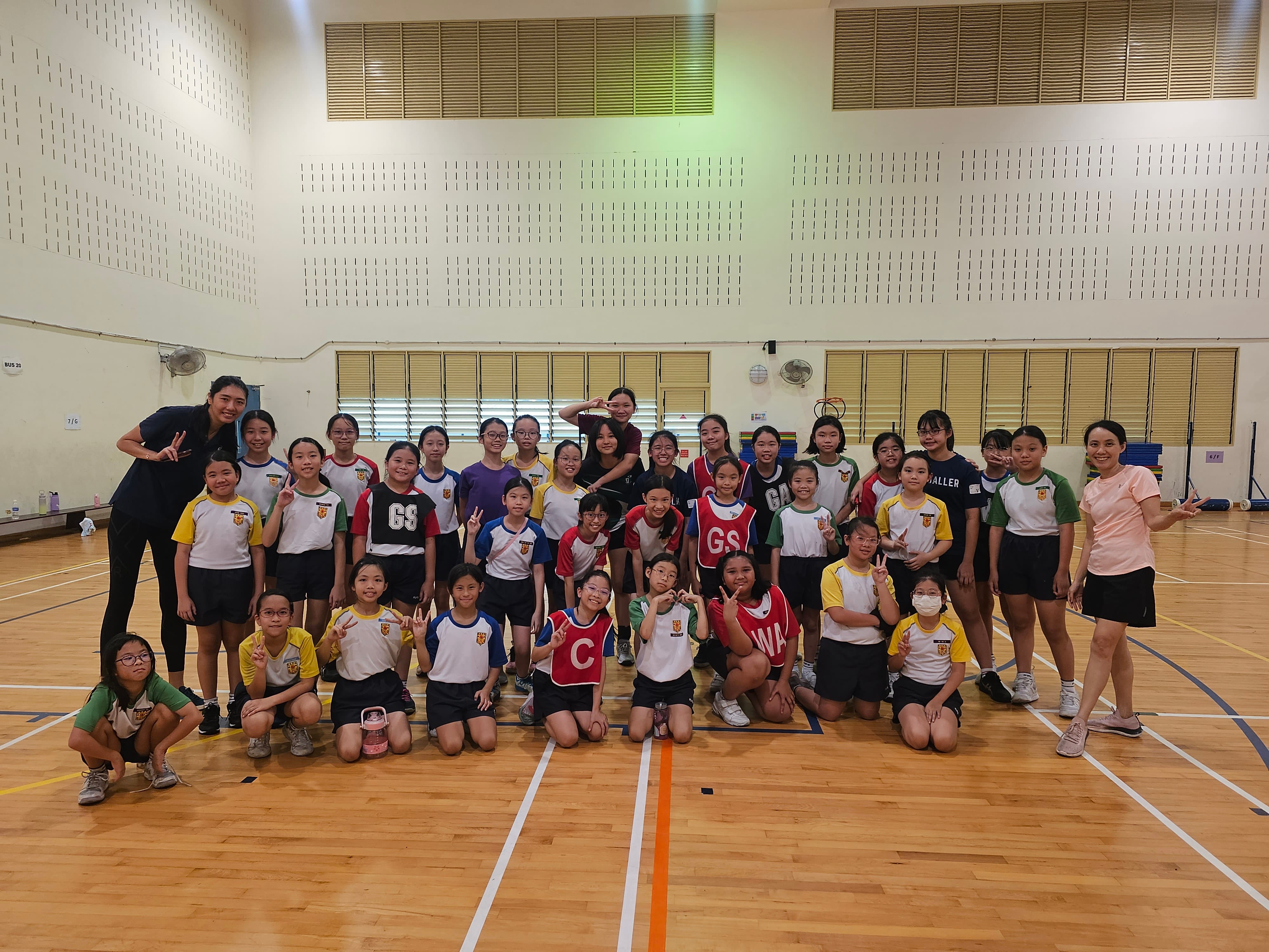 A large group of young girls in sports uniforms pose together inside an indoor gymnasium with wooden flooring.