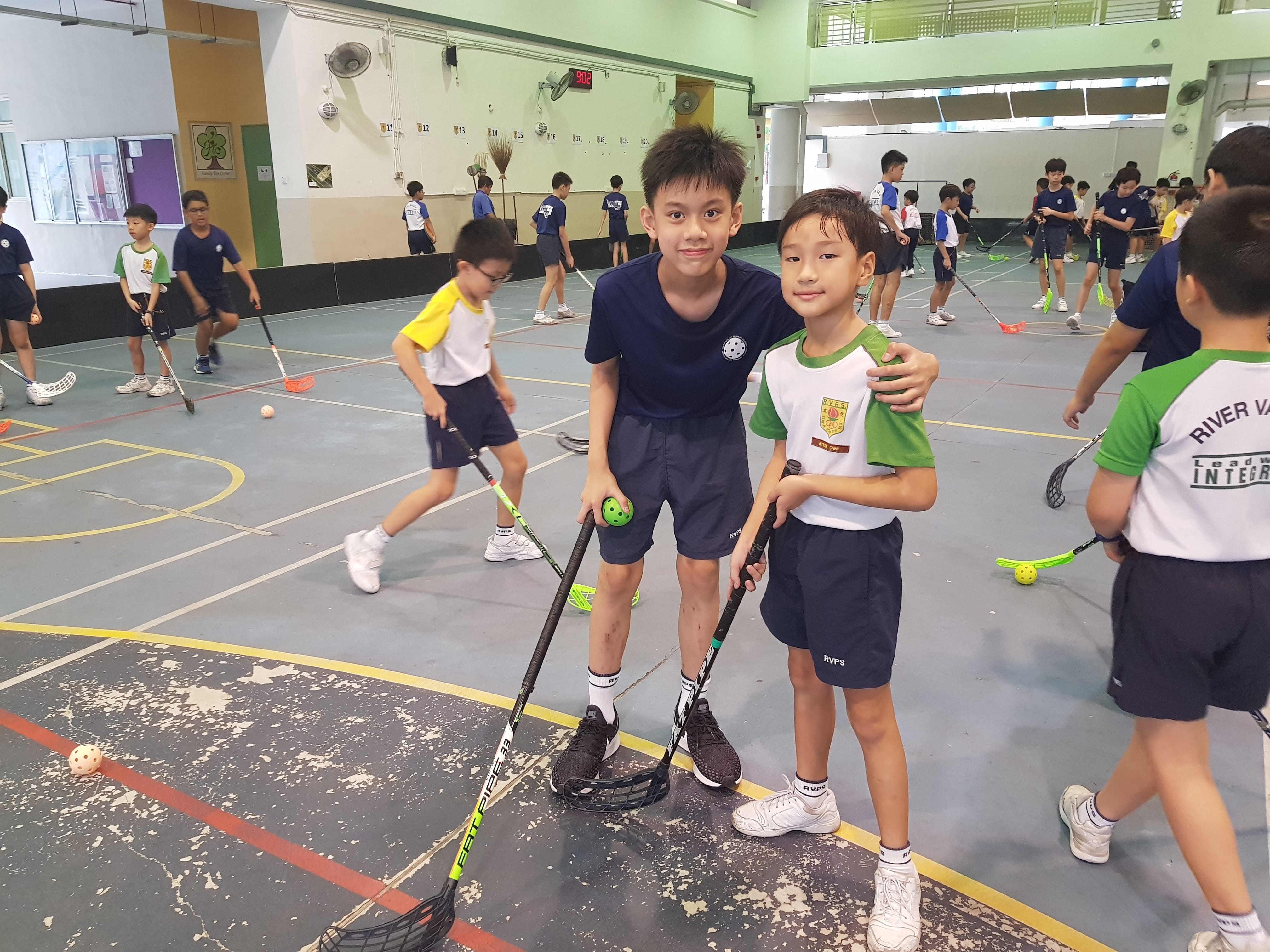 Two boys with floorball sticks pose in a gym while other children practice behind them.