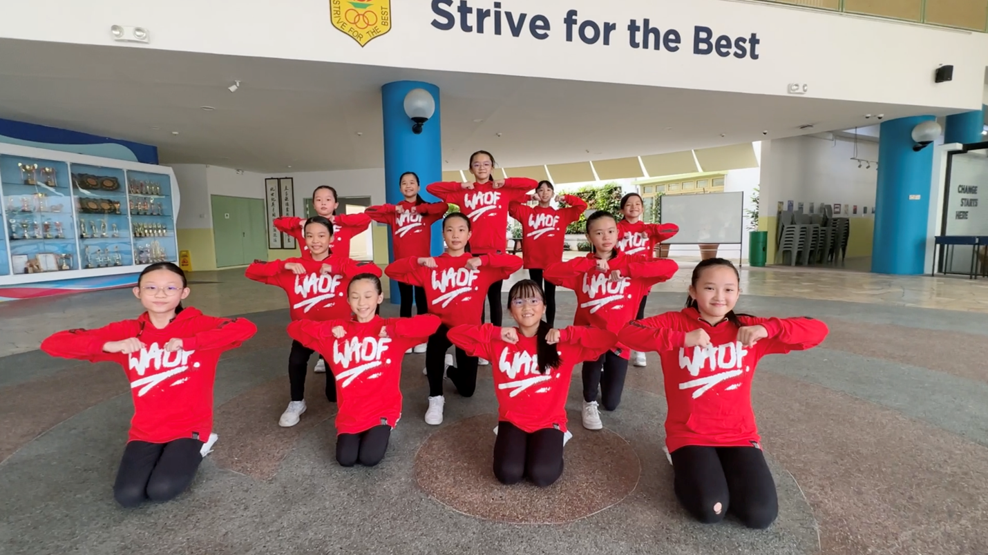 Group of children in red WAOF sweatshirts posing in rows inside a school hall with a Strive for the Best sign.