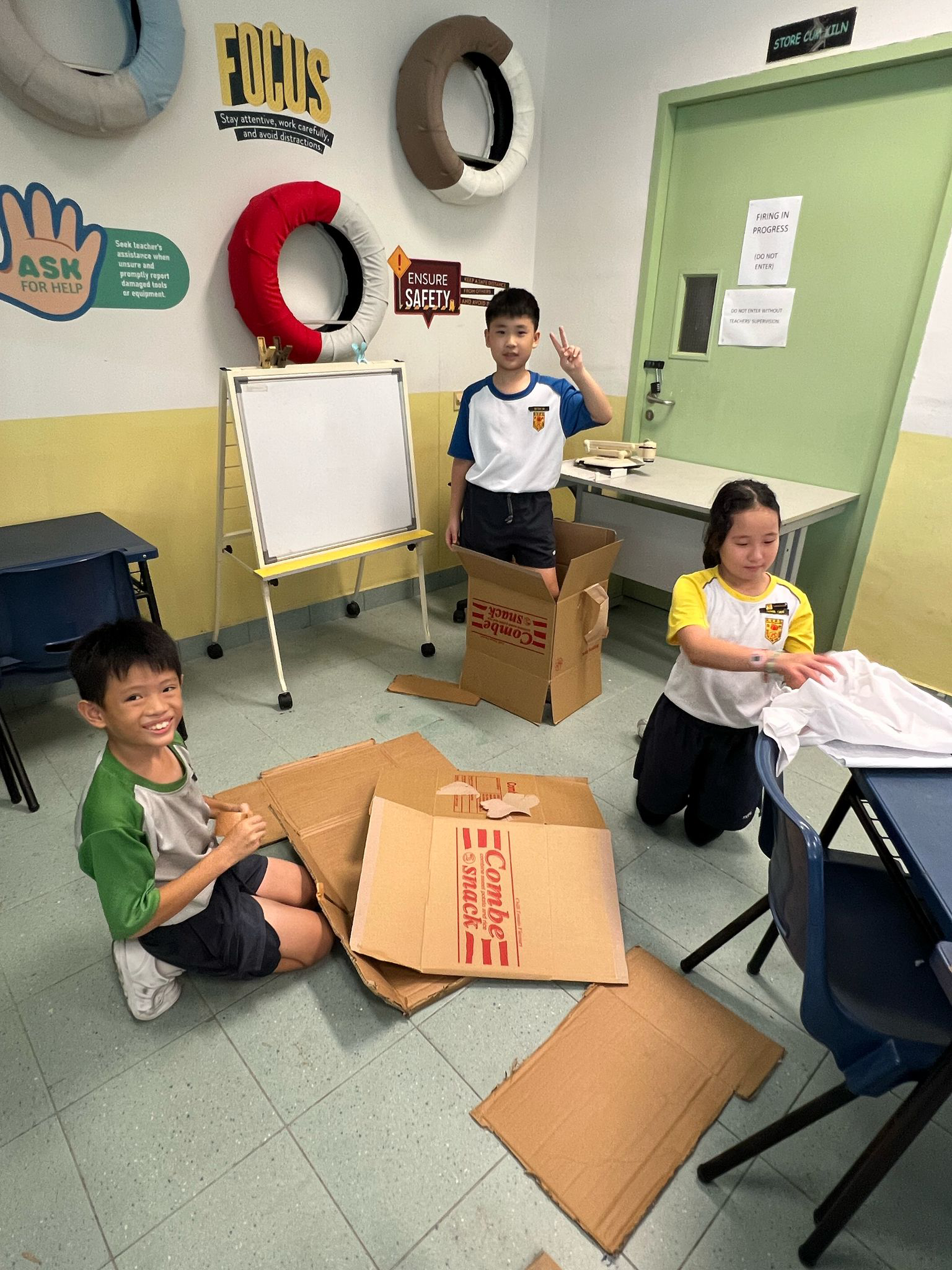 Three children in a classroom with cardboard boxes on the floor; one boy stands in a box making a peace sign.