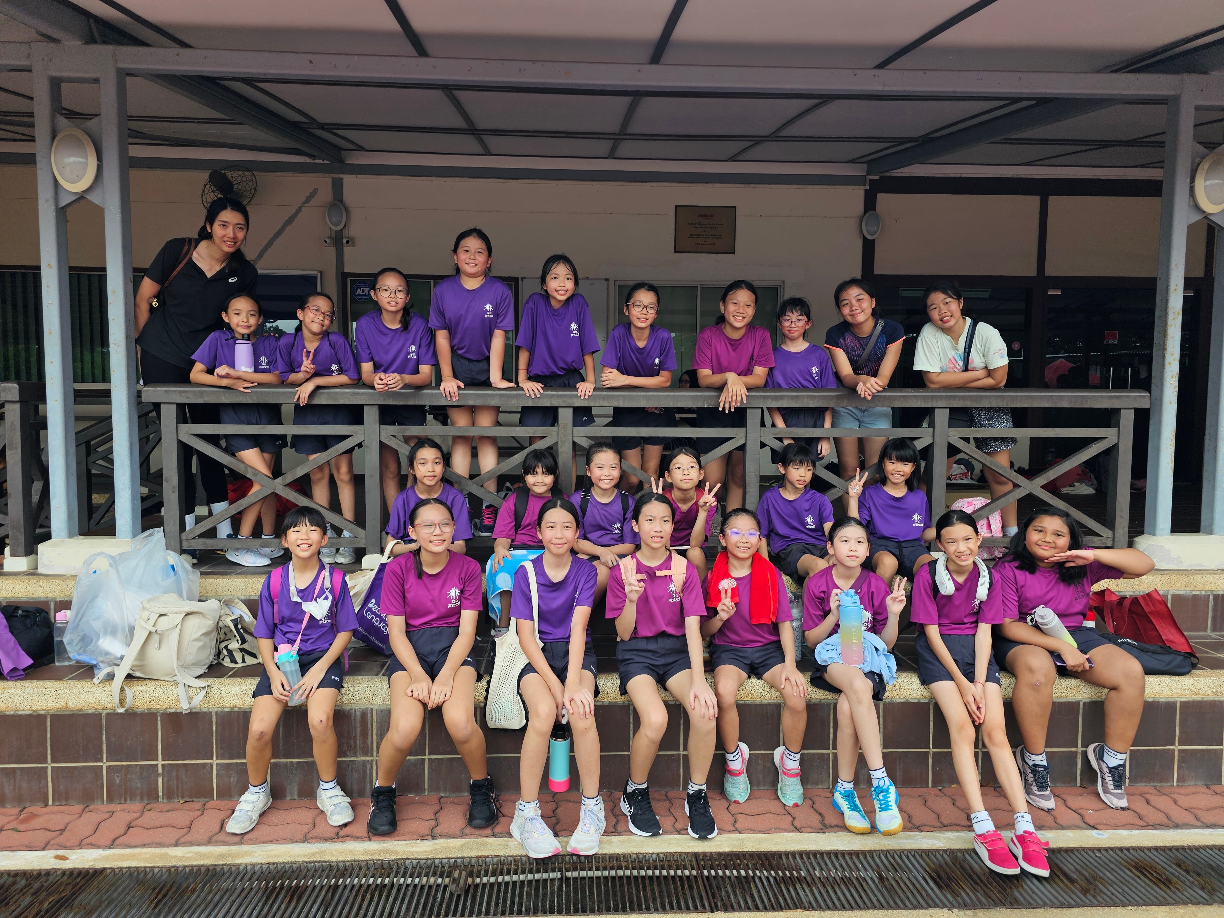 Group of children in purple and pink shirts posing on bleachers, some flashing peace signs.