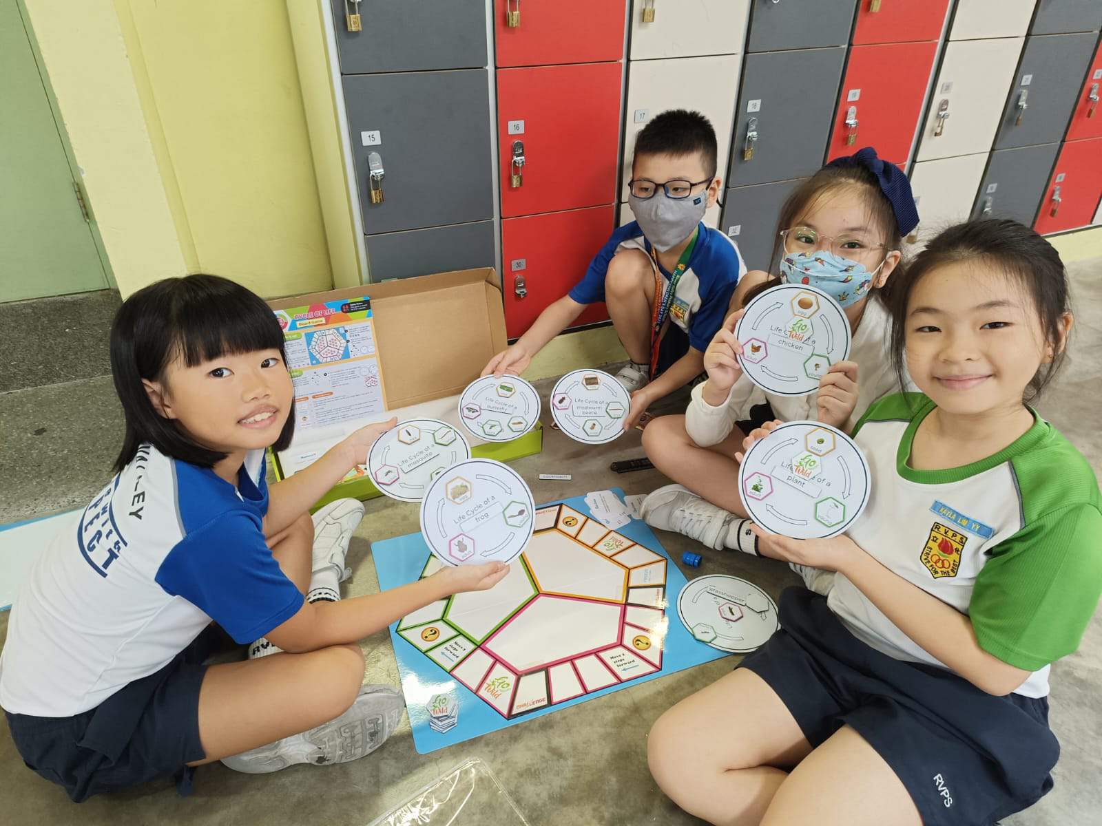 Four children sit on a floor holding circular life cycle diagram cards next to a colorful board game and lockers.