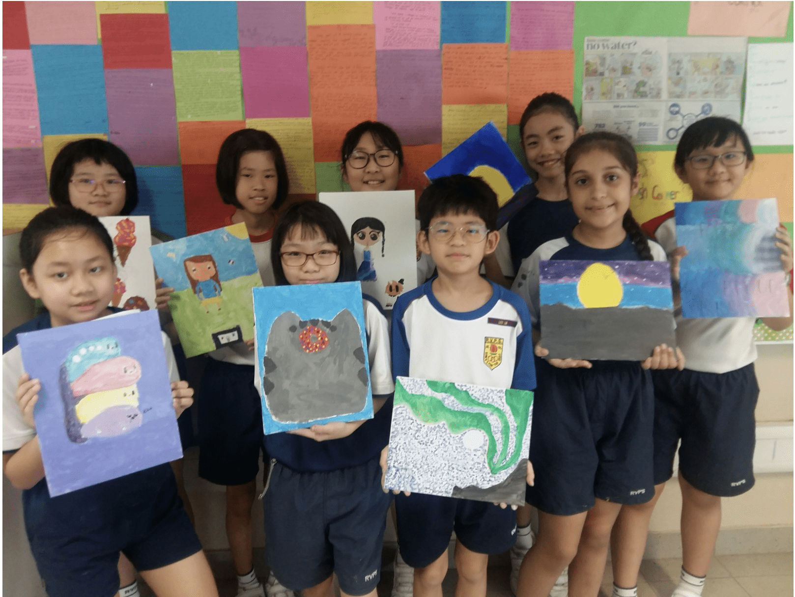 Nine children in school uniforms hold up their colorful painted artworks in front of a multicolored wall display.