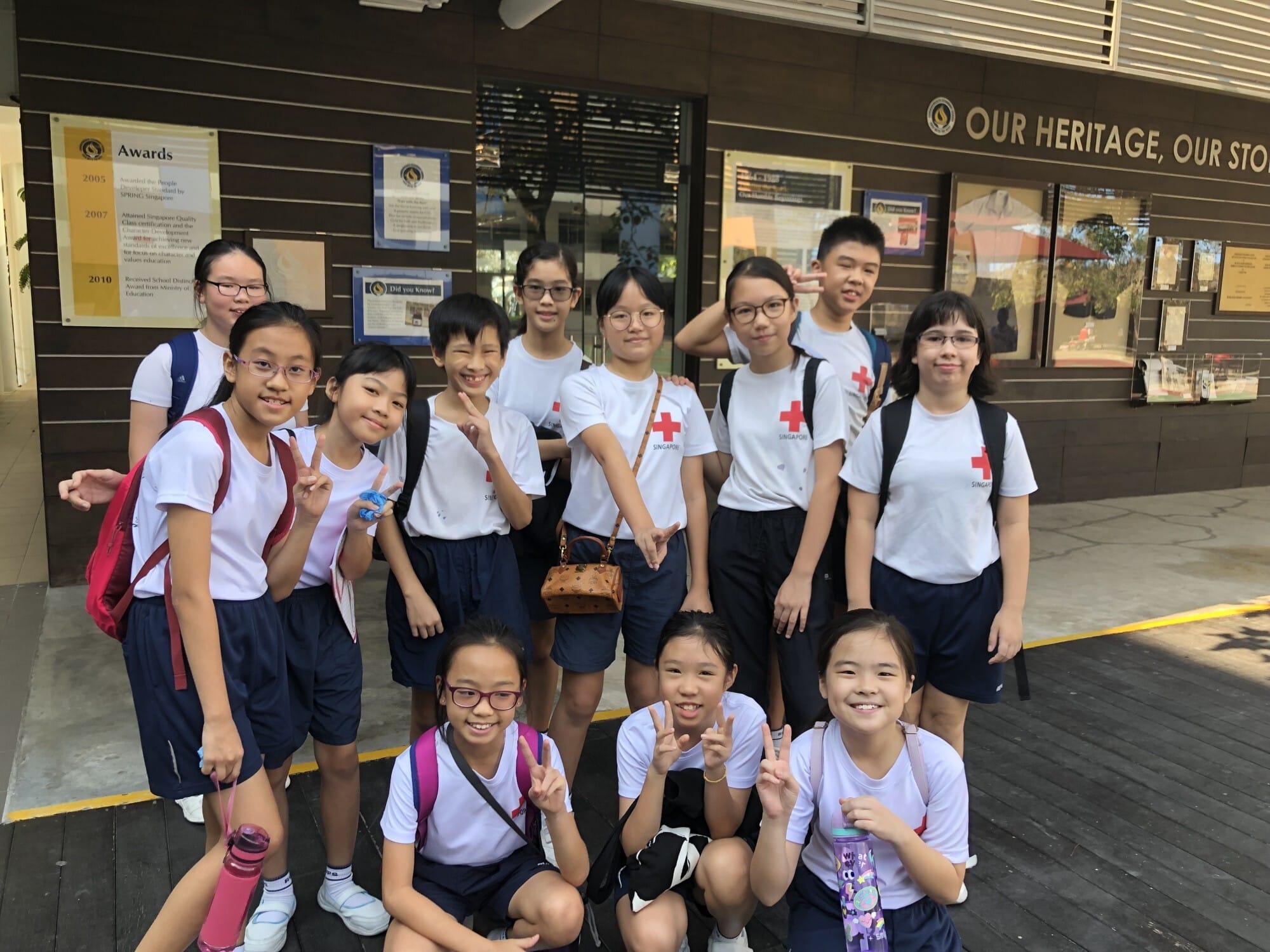 Group of school children in uniforms, some wearing Singapore Red Cross shirts, posing with peace signs outside a school.
