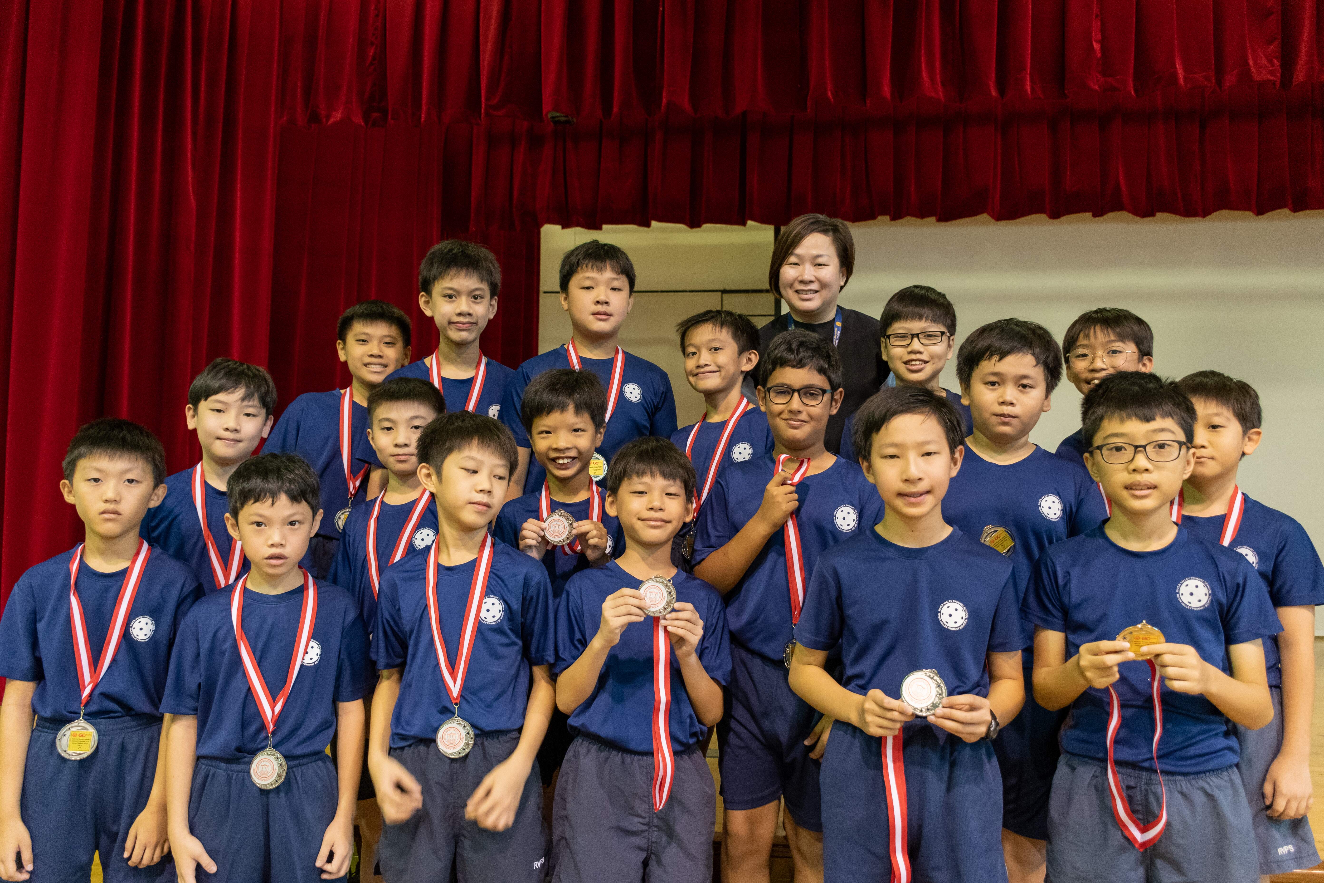 A group of boys in blue uniforms wearing medals pose with a smiling woman in front of a red curtain.