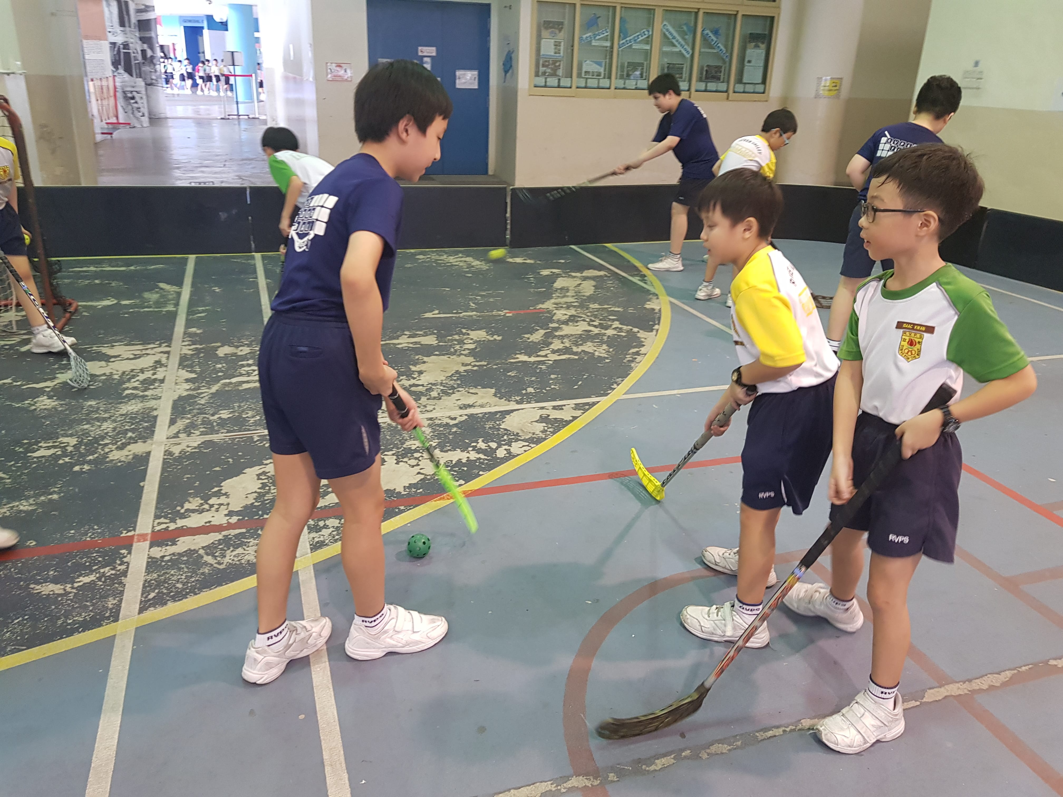 Schoolboys in sports uniforms play floor hockey with sticks and a green ball on an indoor court.