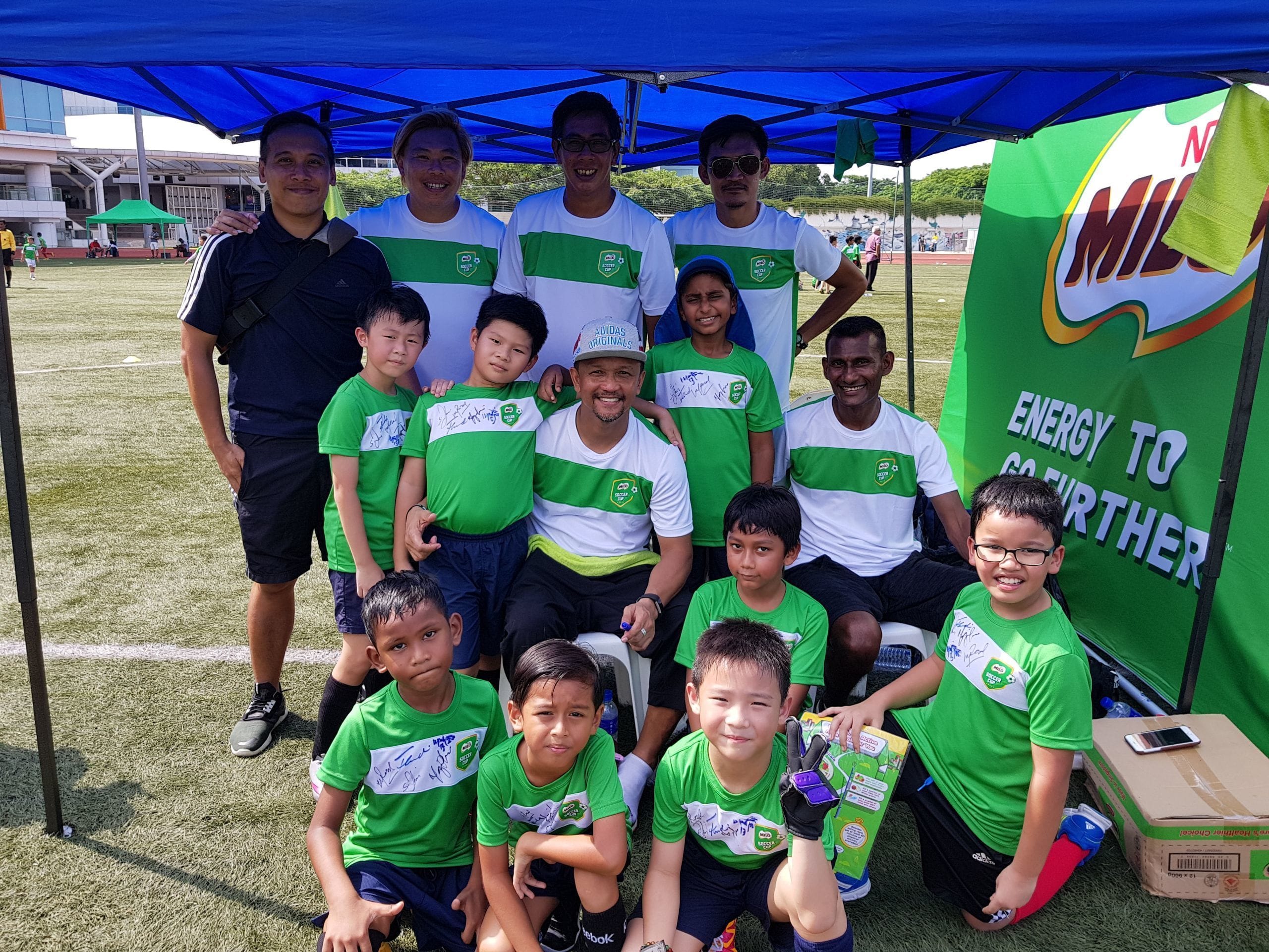Group of adults and children in green soccer jerseys posing under a blue tent next to a Milo banner at a sports field.
