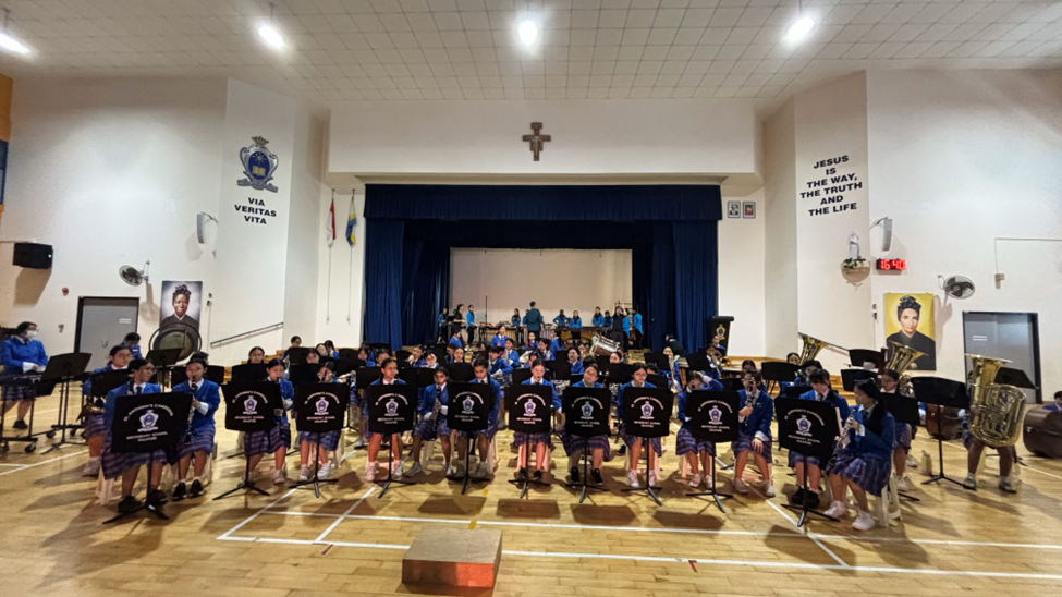 A school concert band performing in a school hall, with students in blue uniforms seated in rows with music stands and various instruments