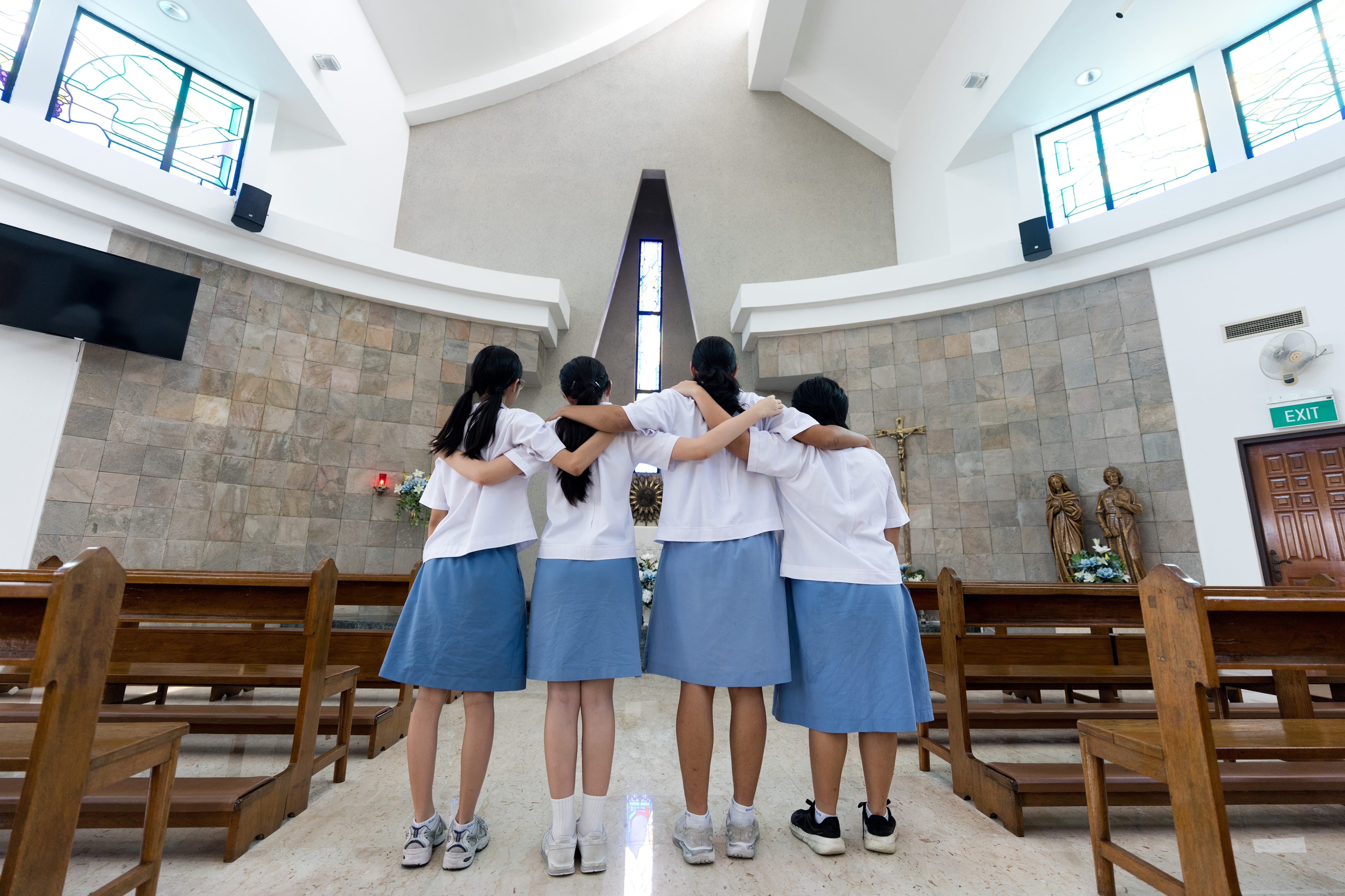 Four students in school uniform standing together with their arms around each other, facing the altar inside a chapel.