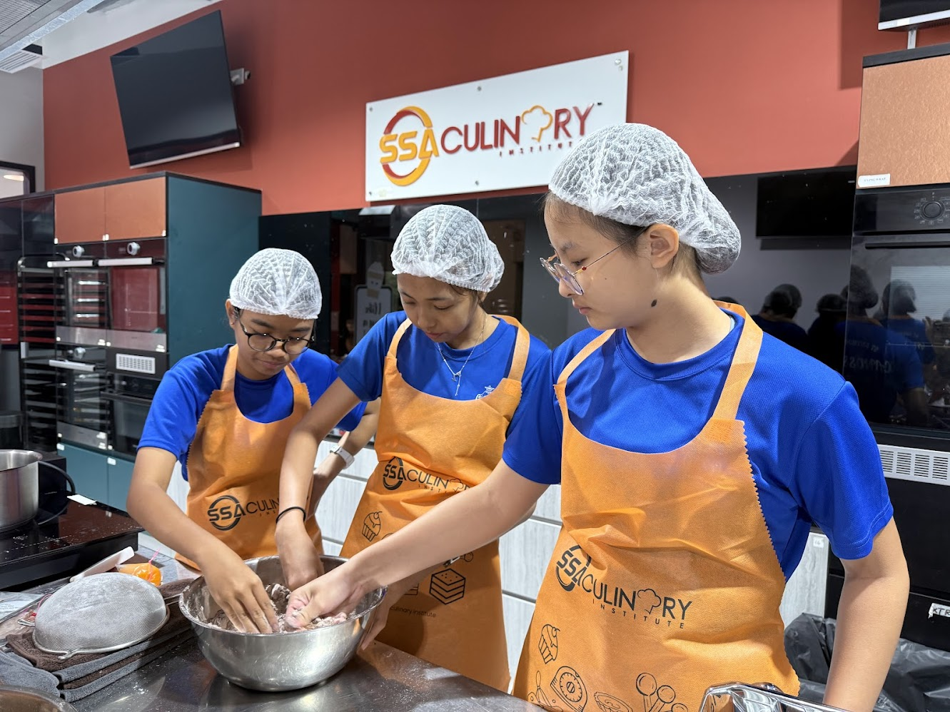 Three students wearing blue T-shirts, orange aprons, and hairnets work together to prepare food in a professional kitchen at SSA Culinary Institute.