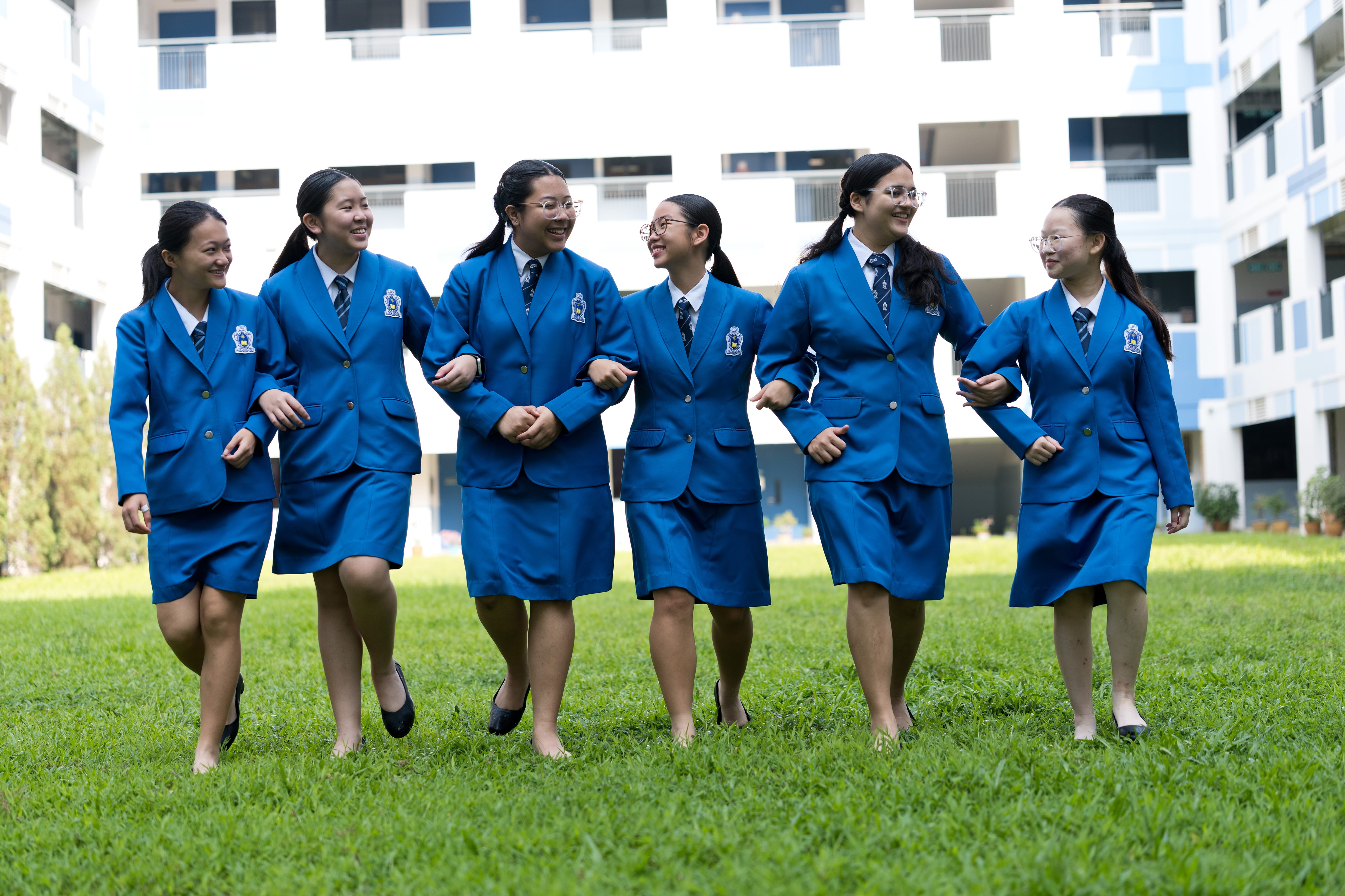 Six students in blue blazer uniforms walking arm in arm across the school field, smiling and laughing together, with the school building in the background.