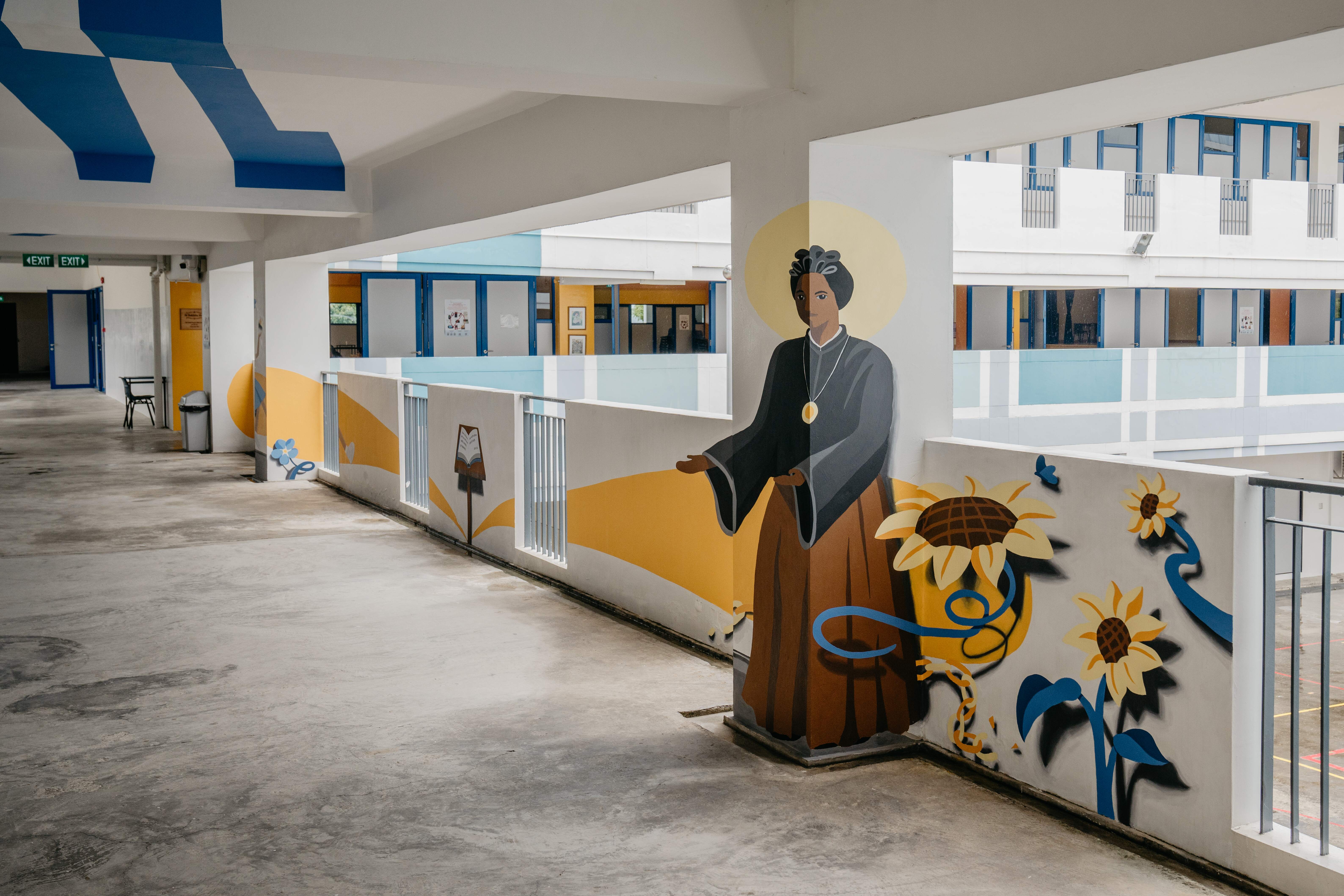 A school corridor with a colorful mural featuring a robed figure with a halo, sunflowers, and blue ribbon designs.
