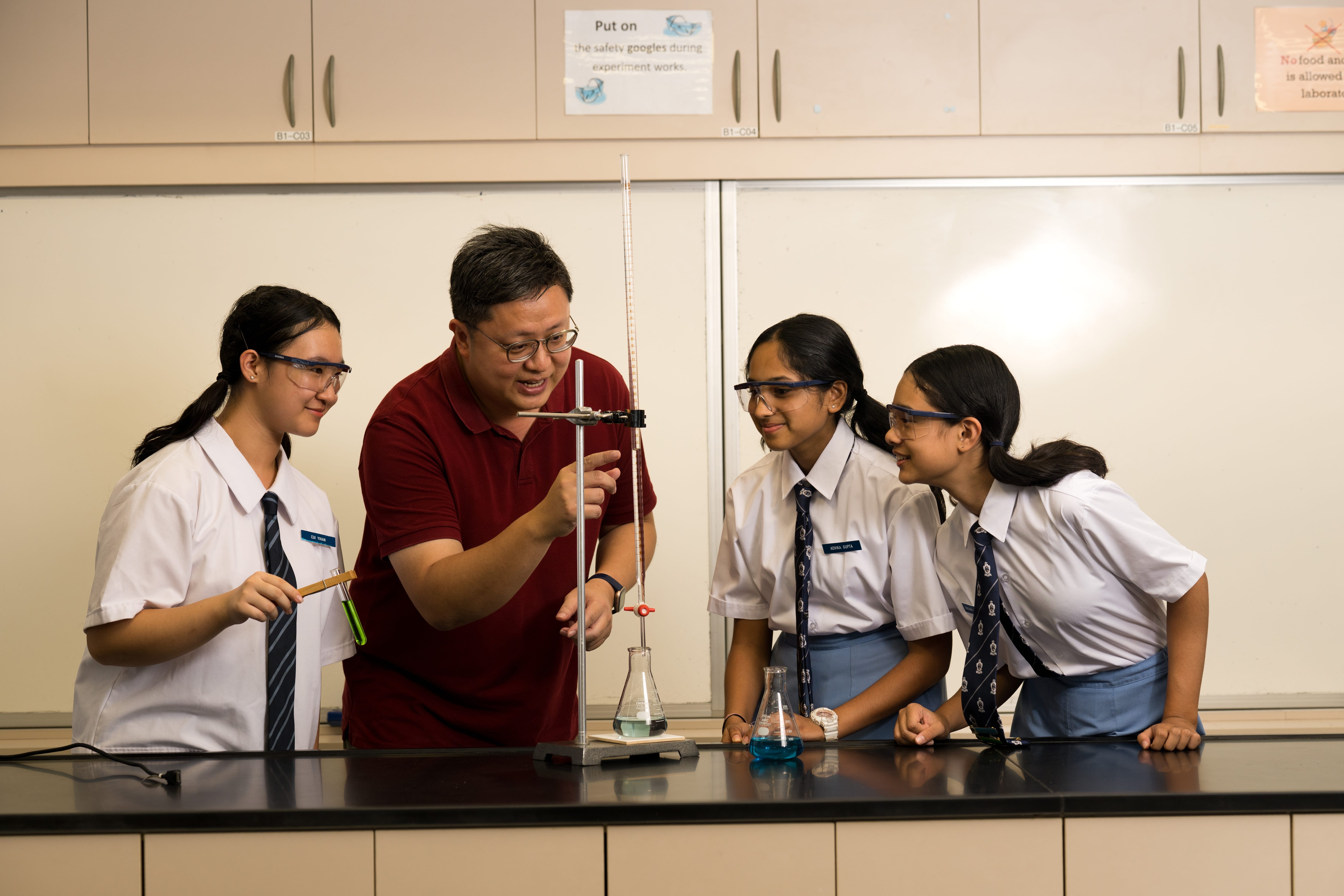 A teacher in a red polo shirt guides three students wearing safety goggles through a science experiment in a laboratory, with a burette, stand, and conical flasks containing blue liquid on the bench in front of them.