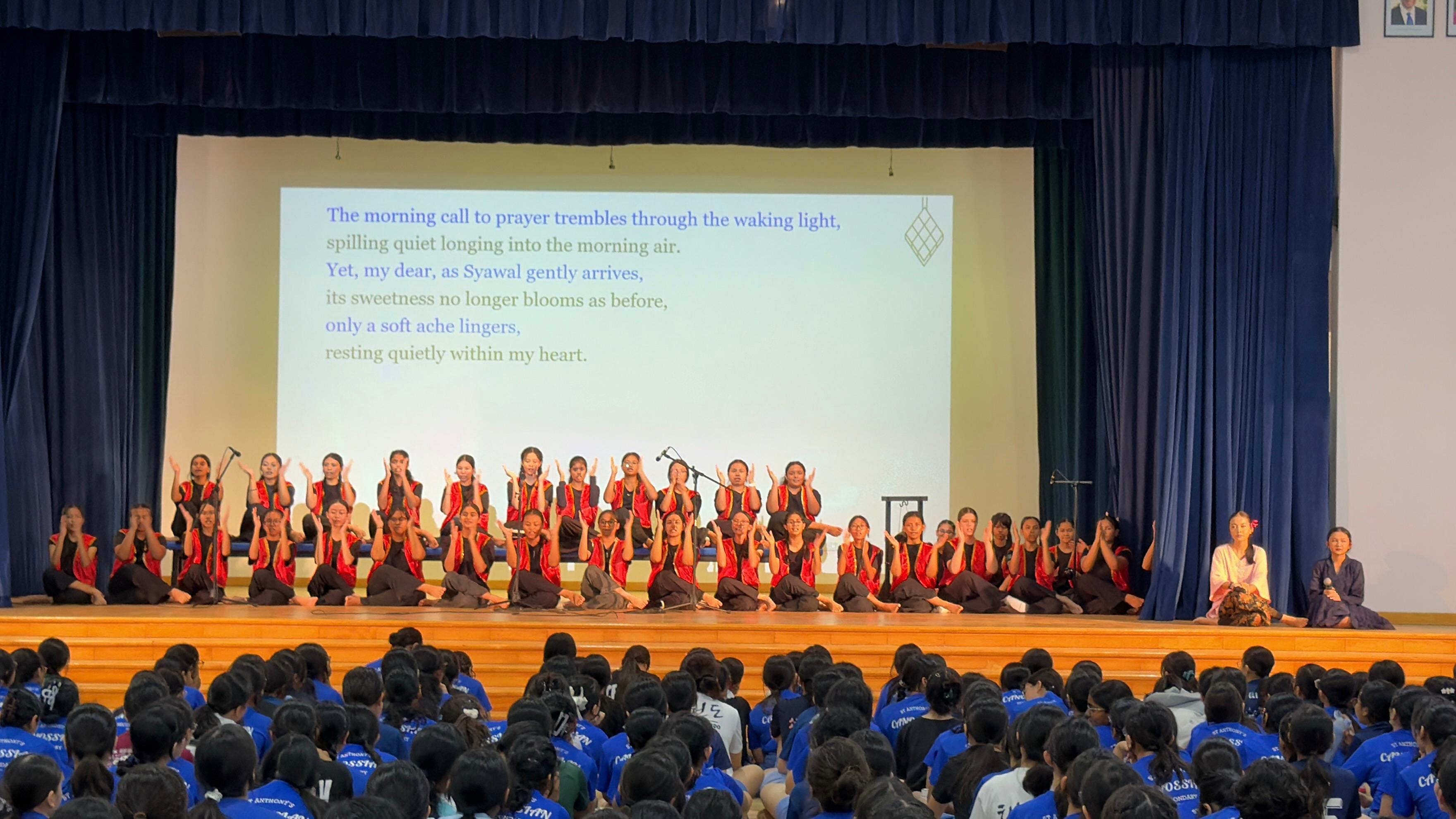 A large group of students in black and red costumes performing a coordinated movement piece on stage, with a projected poem in English about Syawal visible on the screen behind them, in front of an audience of students in blue shirts.