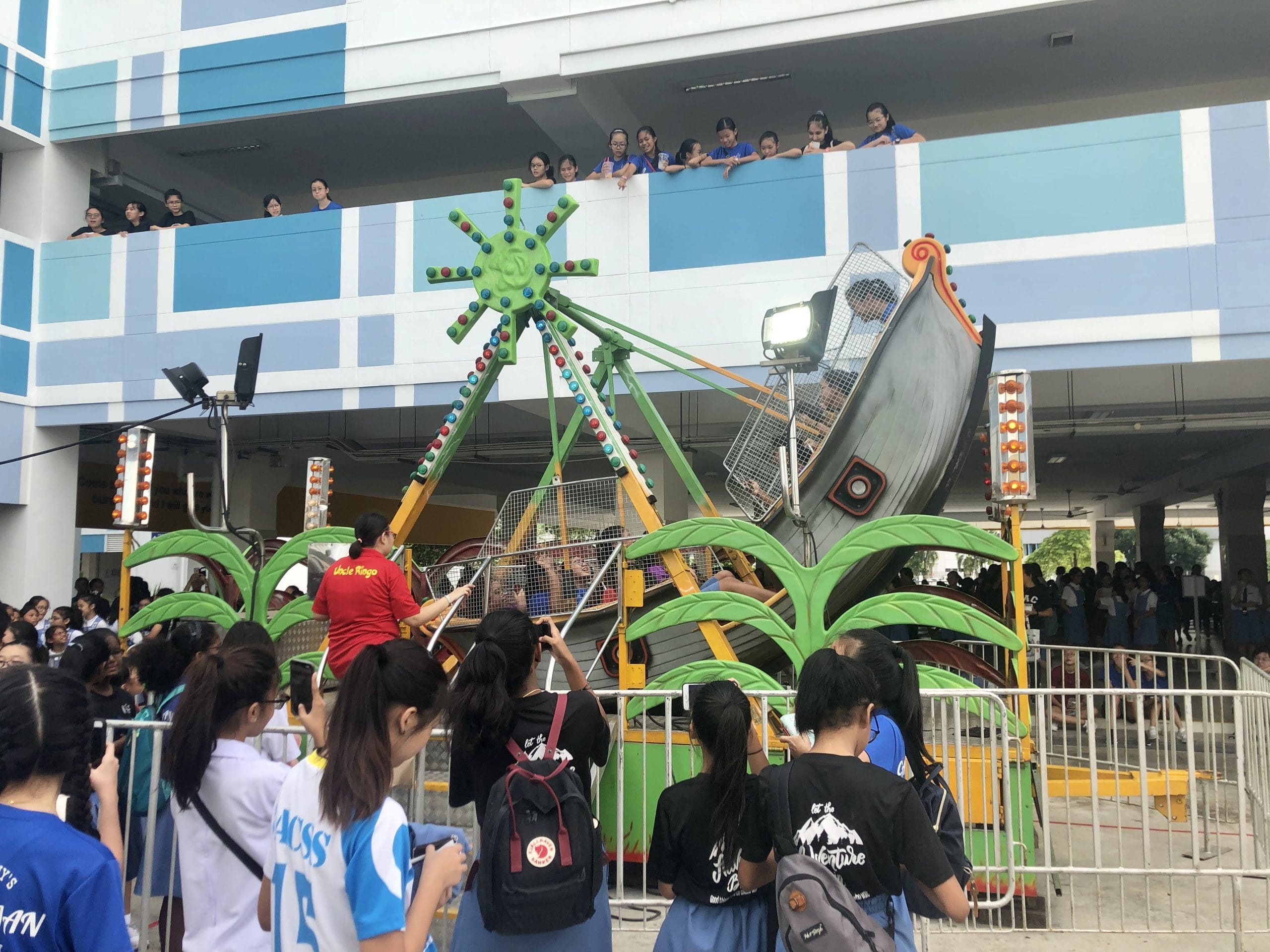 Students crowding around and watching excitedly as a pirate ship carnival ride operates in the school compound, with more students looking on from the upper floor balcony during a school carnival event.