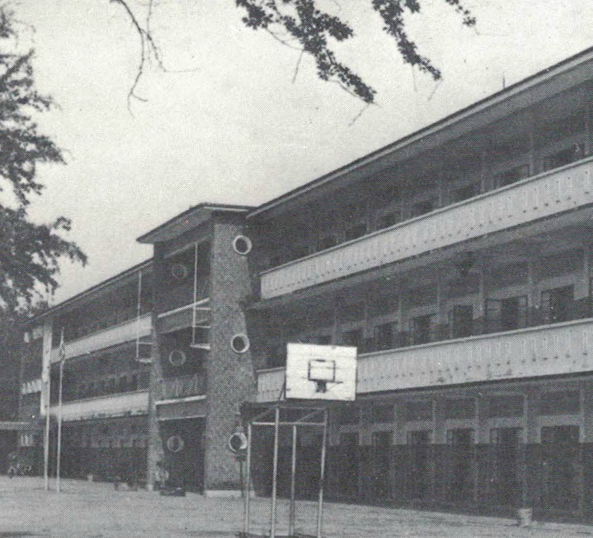 Black and white photo of a two-story school building with a basketball hoop in the foreground and trees nearby.