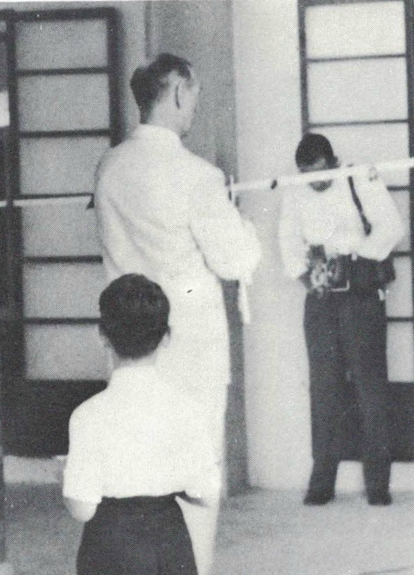 Two people in white uniforms practice a martial arts staff technique while a third person watches in a dojo.