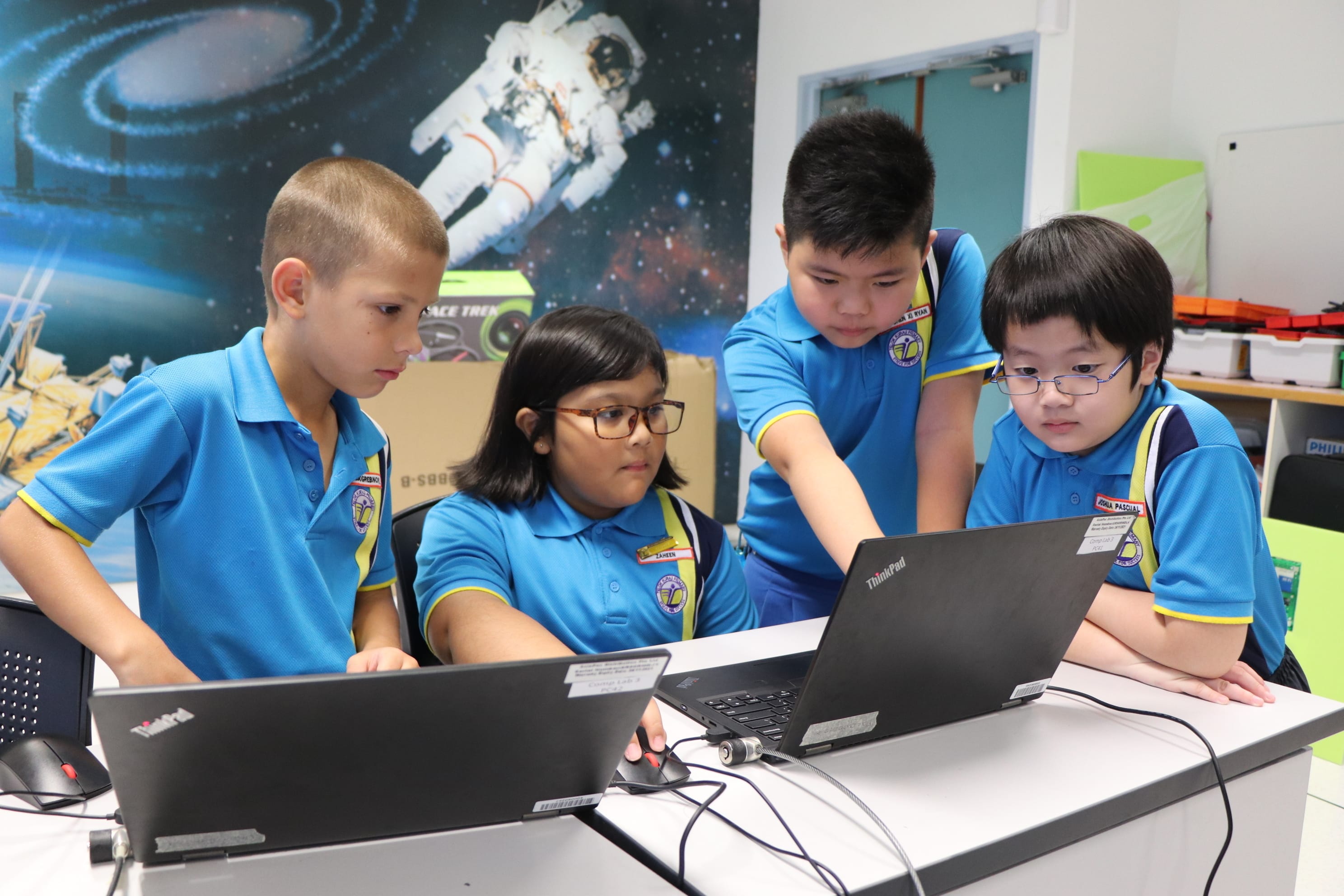 Four children in blue school uniforms gather around ThinkPad laptops in a classroom with a space-themed mural.