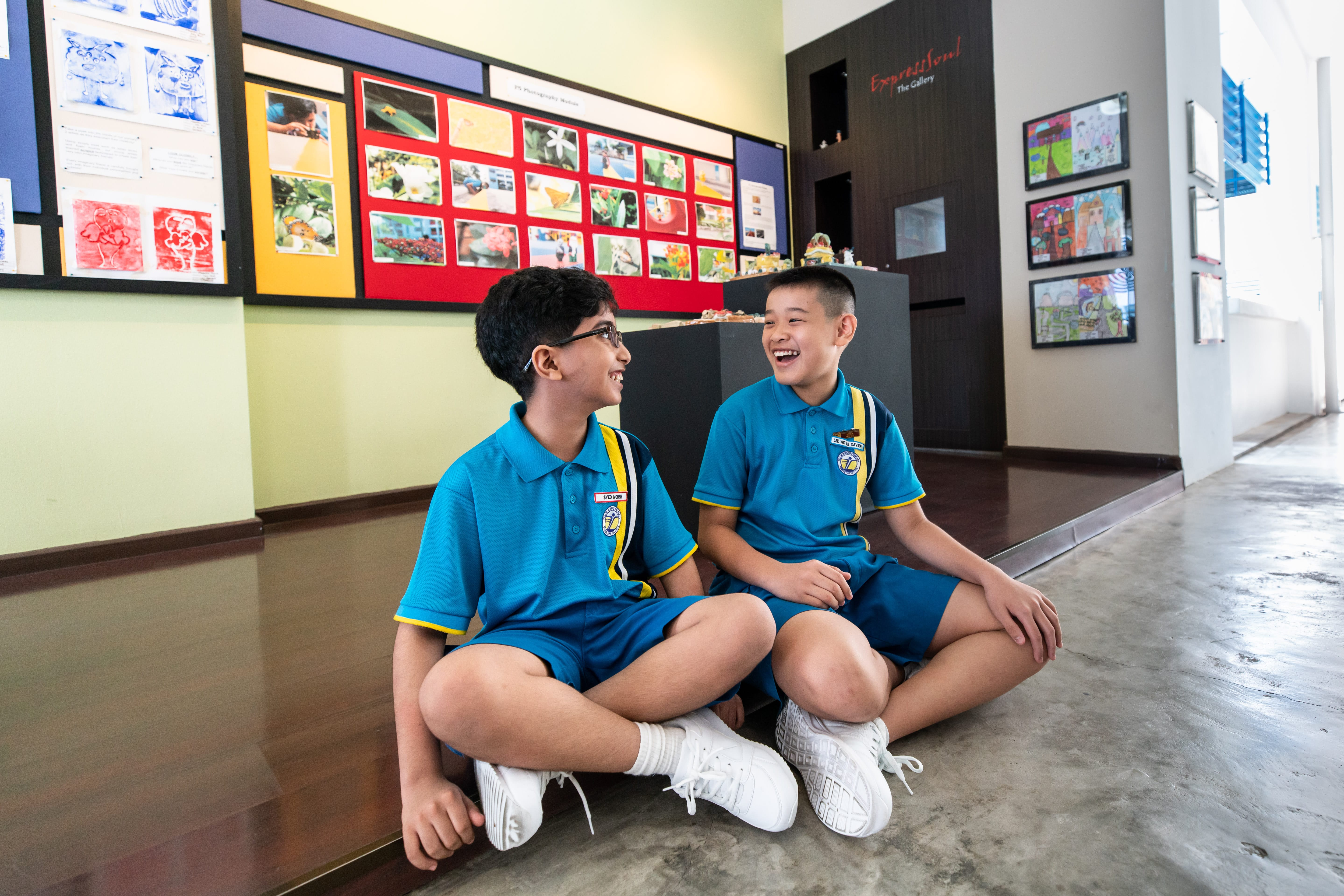 Two boys in blue school uniforms sit on the floor laughing in a school gallery with colorful artwork on the walls.