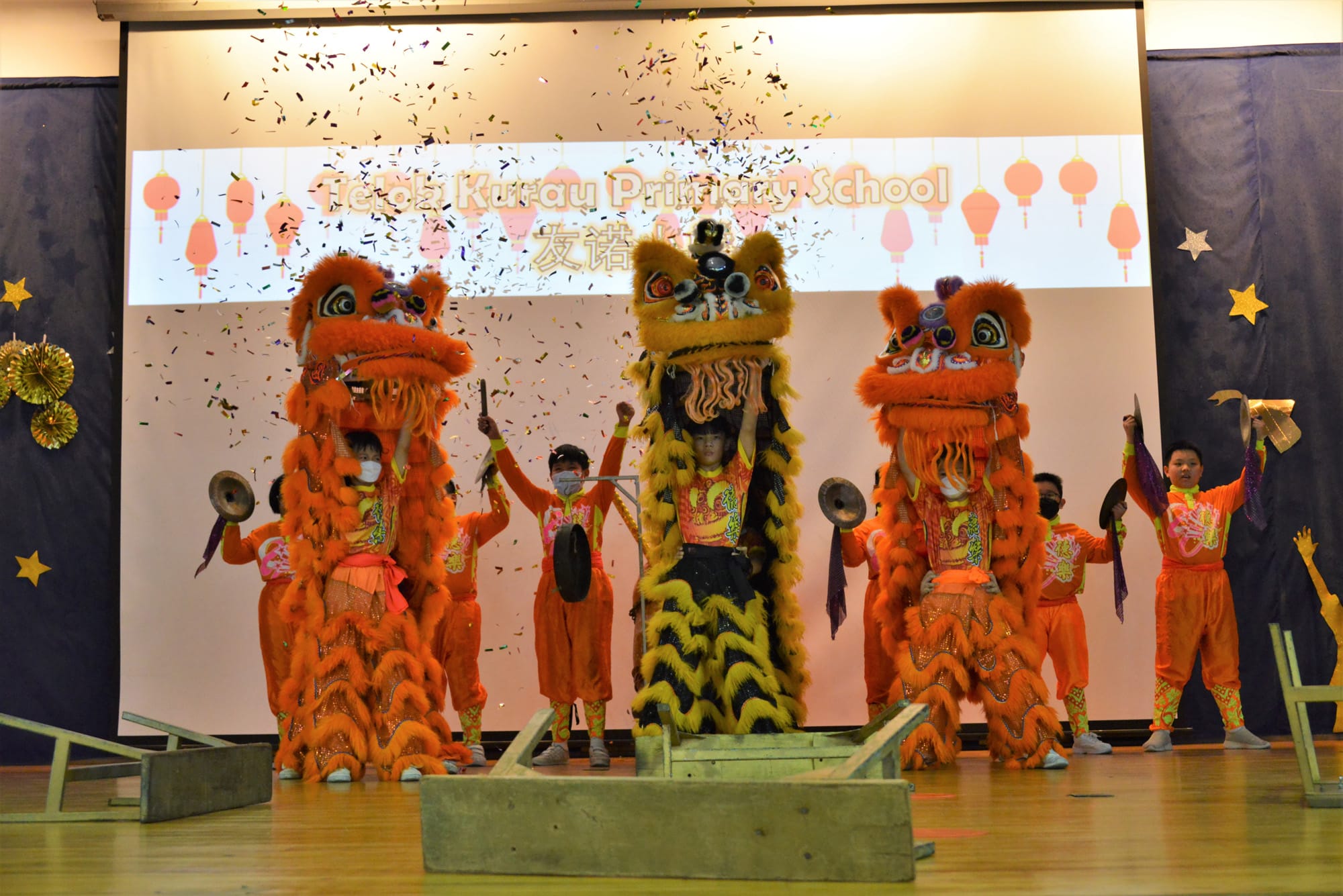 Three colorful lion dance costumes performed on stage with confetti falling at Telok Kurau Primary School.