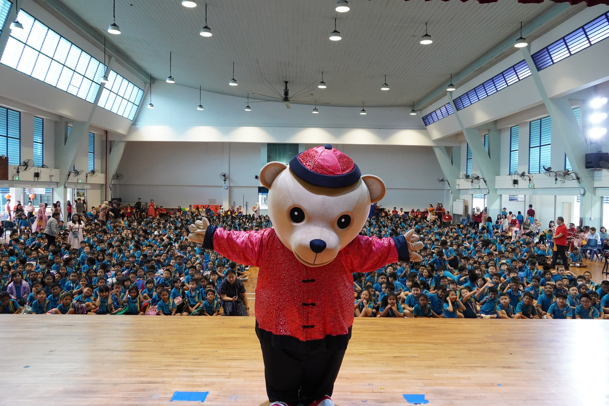A bear mascot in a red traditional outfit stands on stage before a large crowd of children in blue uniforms in a school hall.