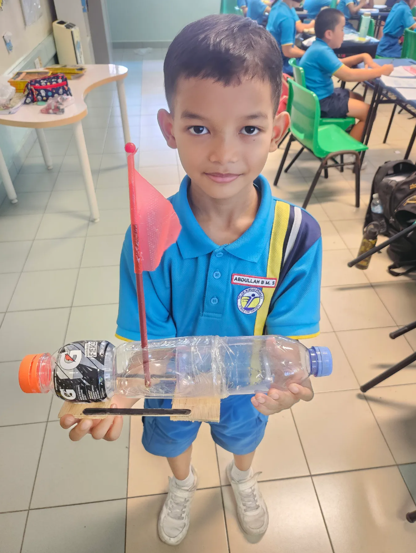 A young boy in a blue school uniform holds a handmade boat craft using a plastic bottle with a small red flag.