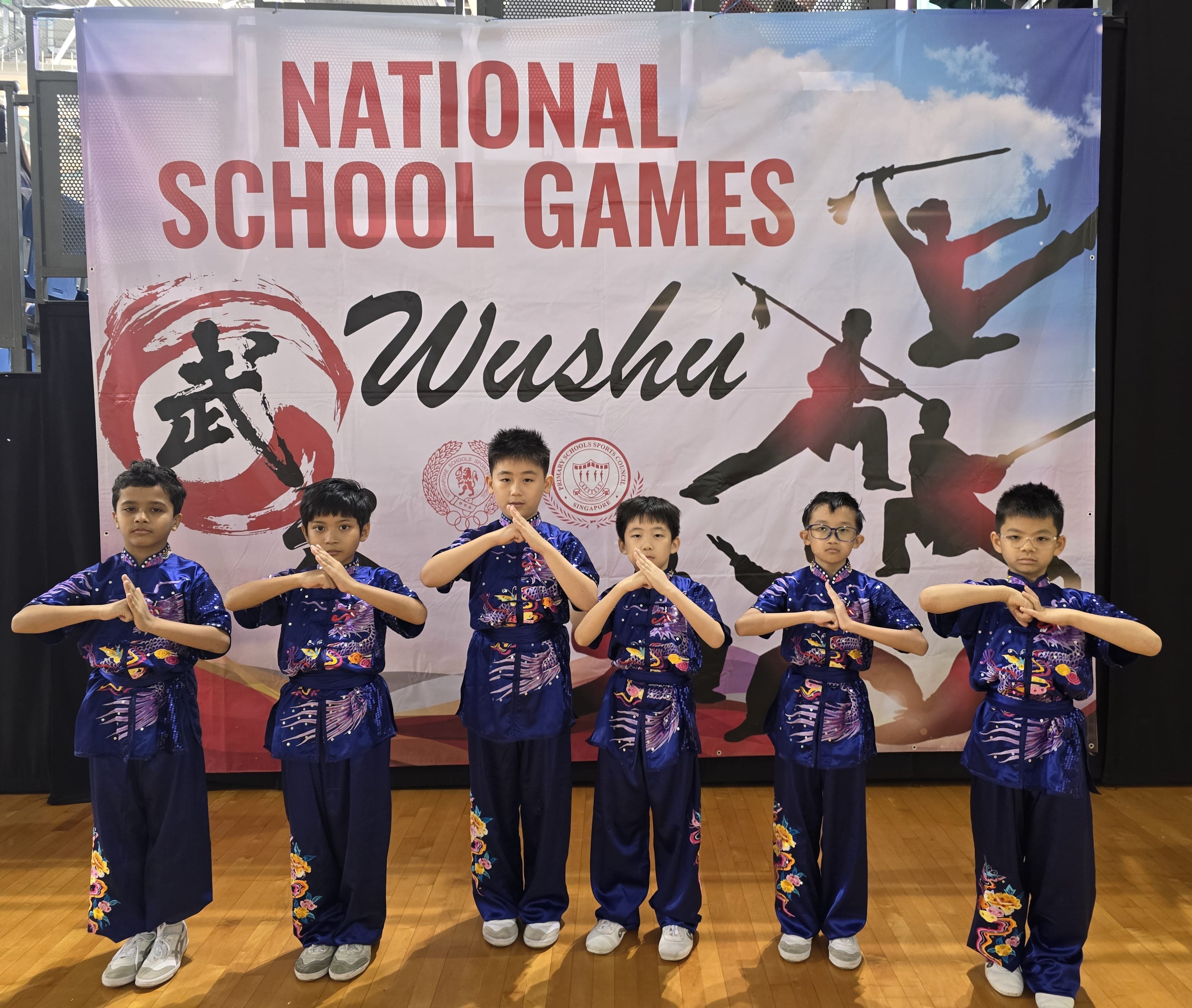 Six children in blue wushu uniforms pose with clasped hands before a National School Games Wushu banner.