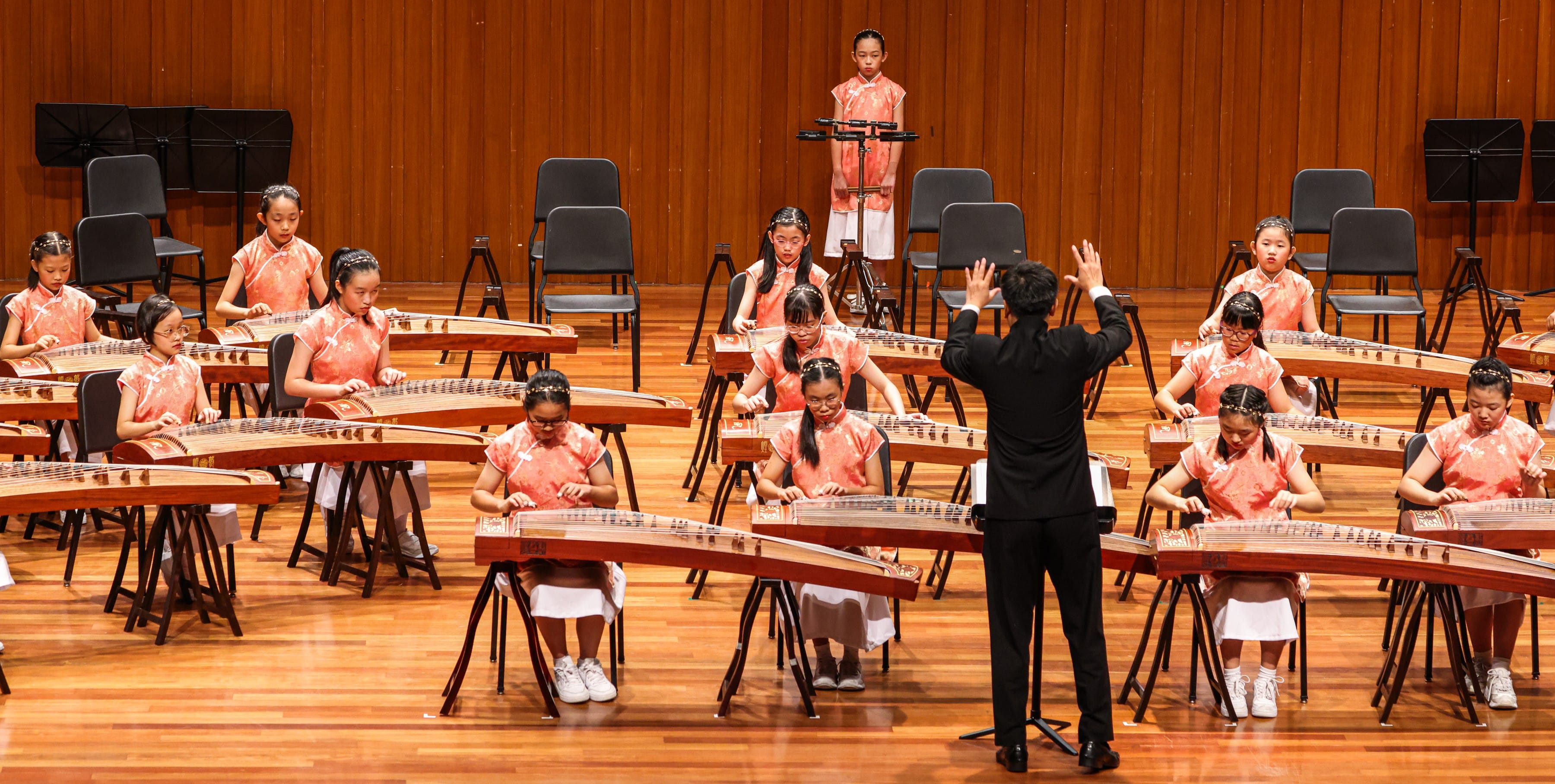 Children in pink qipao play guzheng instruments on a wooden stage while a conductor in black leads them.