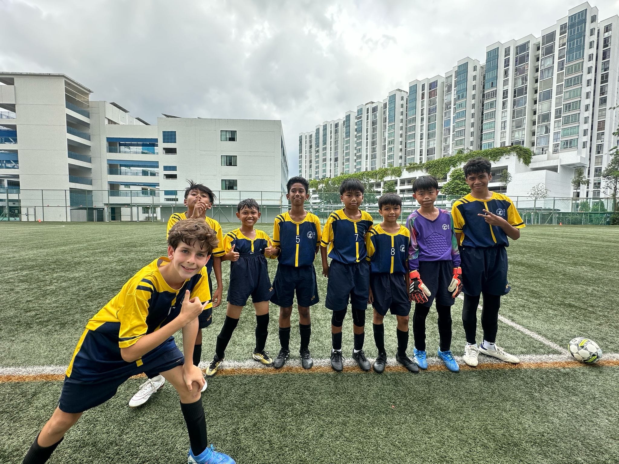 Eight young boys in yellow and navy soccer uniforms pose on a green field with urban buildings in the background.