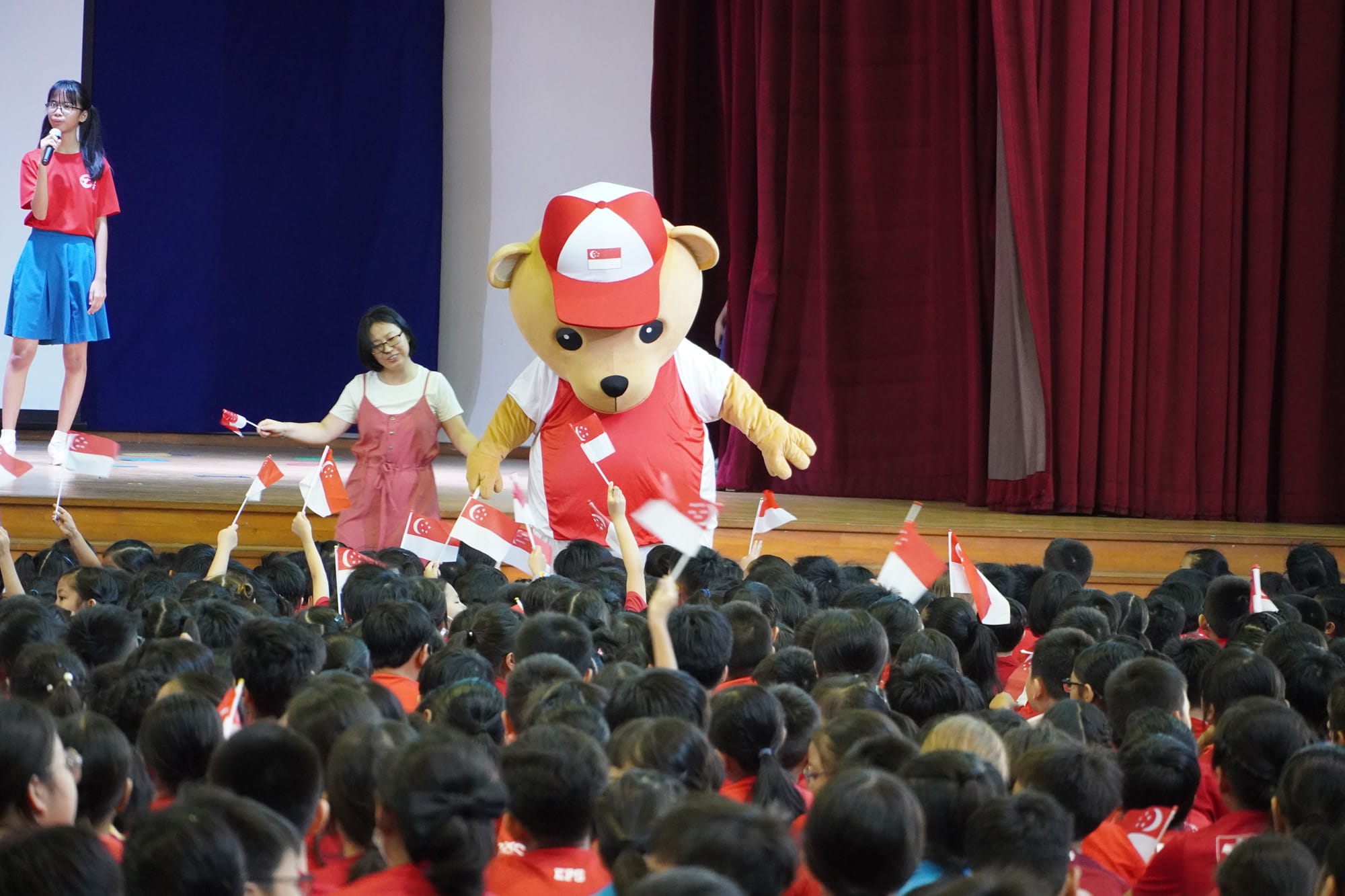 A bear mascot in Singapore jersey and cap stands on stage as children wave Singapore flags below.