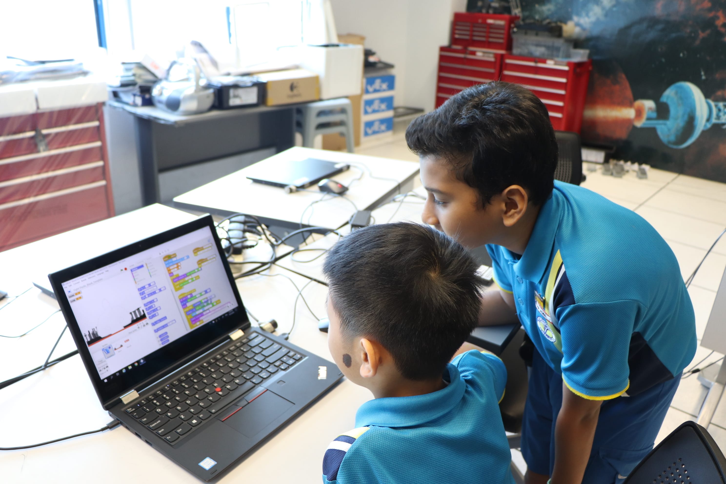 Two boys in blue shirts look at a laptop displaying a Scratch coding project on a desk.