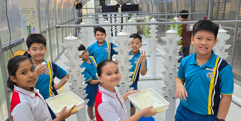 Six children in school uniforms smile near tall white vertical hydroponic towers inside a greenhouse.
