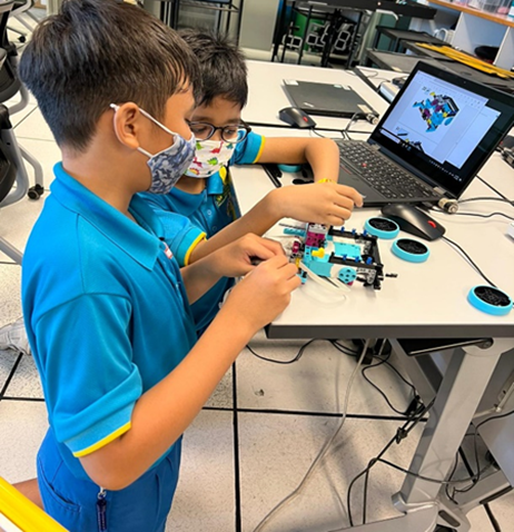 Two masked boys in blue uniforms build a colorful LEGO robot at a desk with a laptop showing a 3D model.
