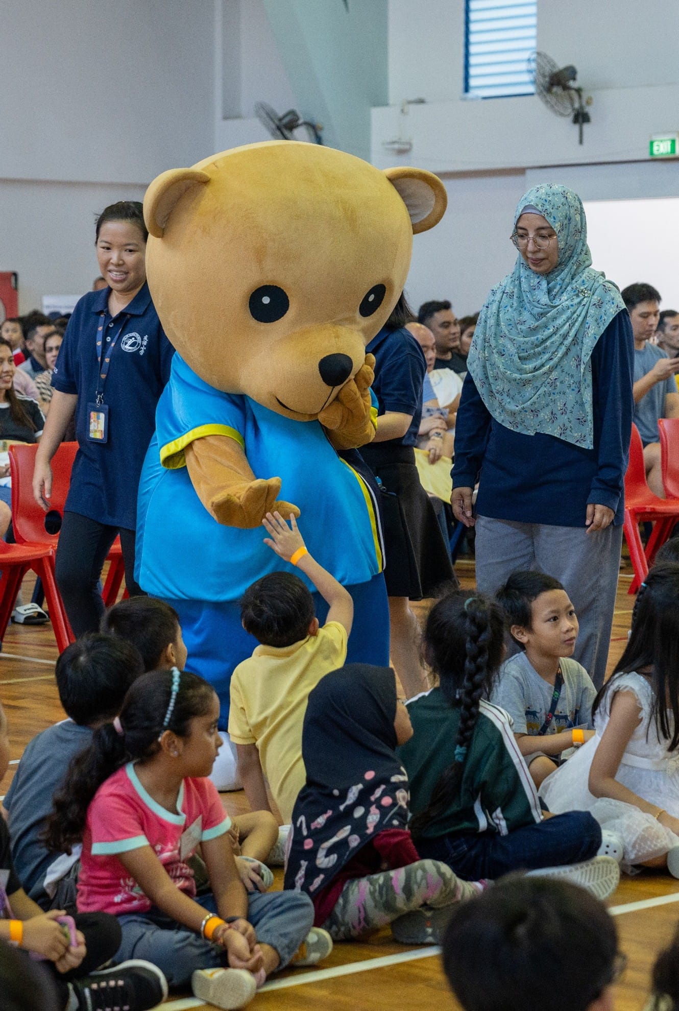 A large bear mascot in a blue shirt interacts with children seated on a school hall floor, with adults standing nearby.