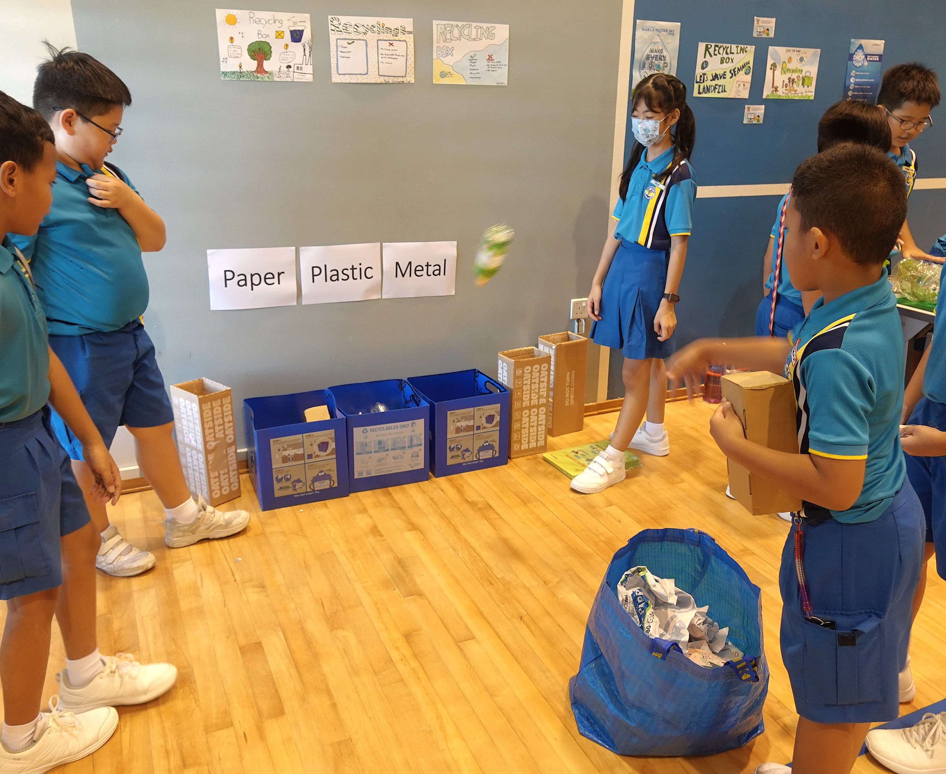 Students in blue uniforms sort recyclables into bins labeled Paper, Plastic, and Metal in a classroom.
