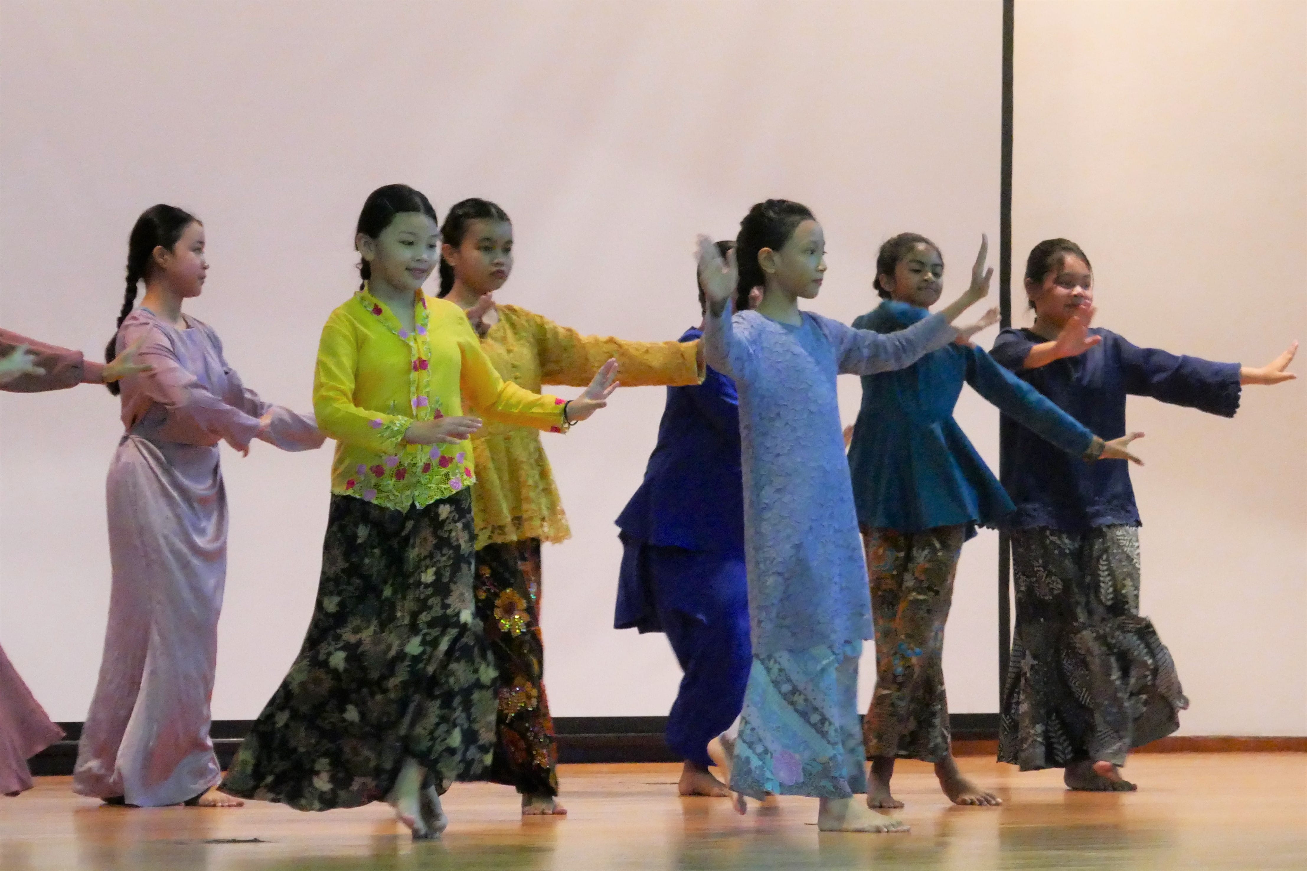 Six girls in colorful traditional costumes perform a coordinated dance on a wooden stage with arms extended.