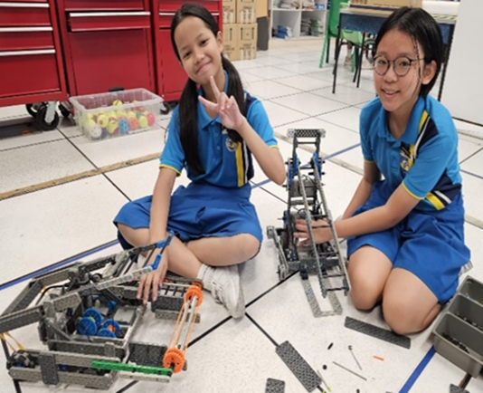 Two girls in blue uniforms sit on a tiled floor with assembled robotics projects between them.