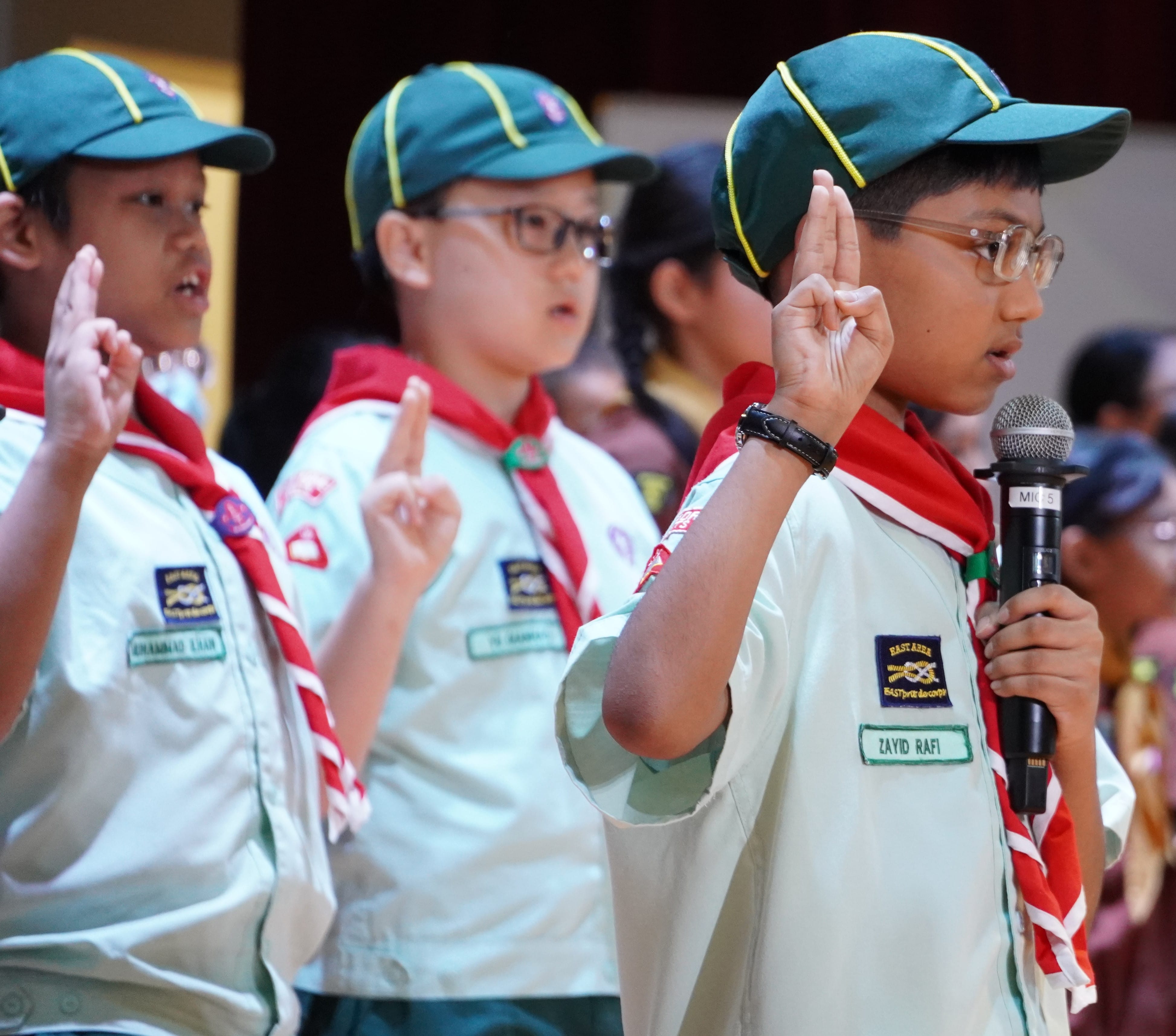 Three young scouts in green caps and red scarves raise their hands in a pledge, with one boy holding a microphone.