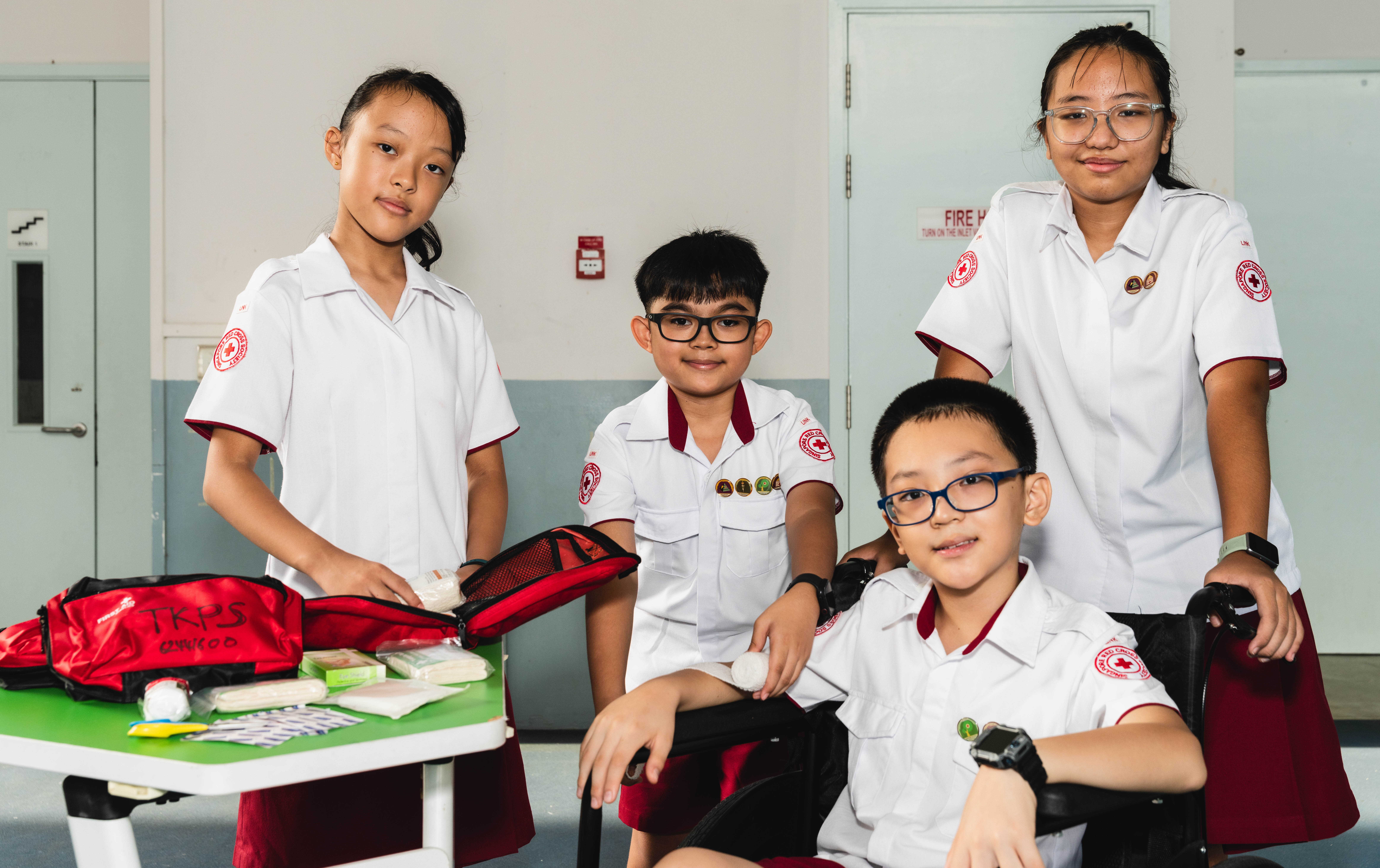 Four youths in white uniforms pose by a table with a red first-aid kit and bandages; one sits in a wheelchair.