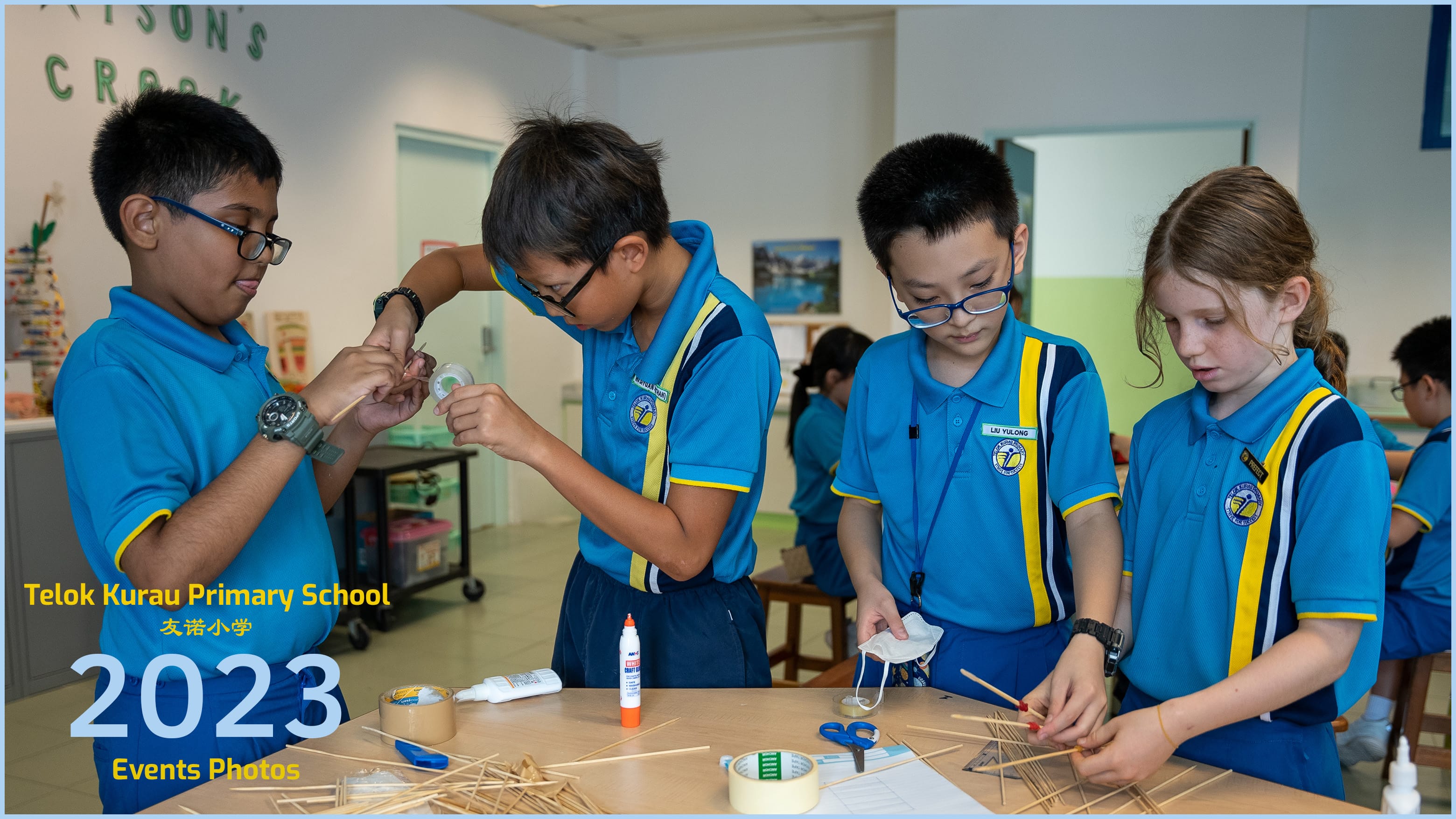 Four primary school students in blue uniforms build a structure using wooden sticks, glue, and tape at a classroom table.