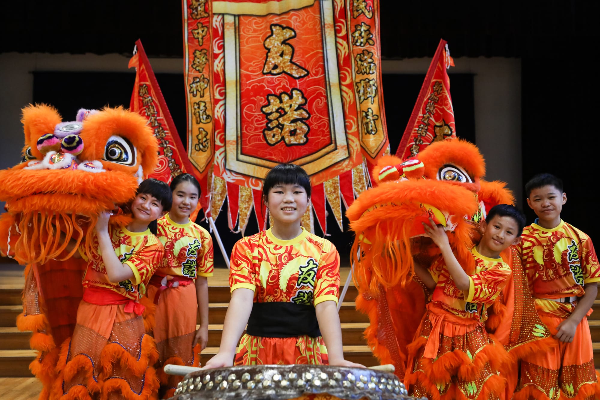 Children in orange and yellow costumes pose with two orange lion dance heads and red banners with Chinese characters.